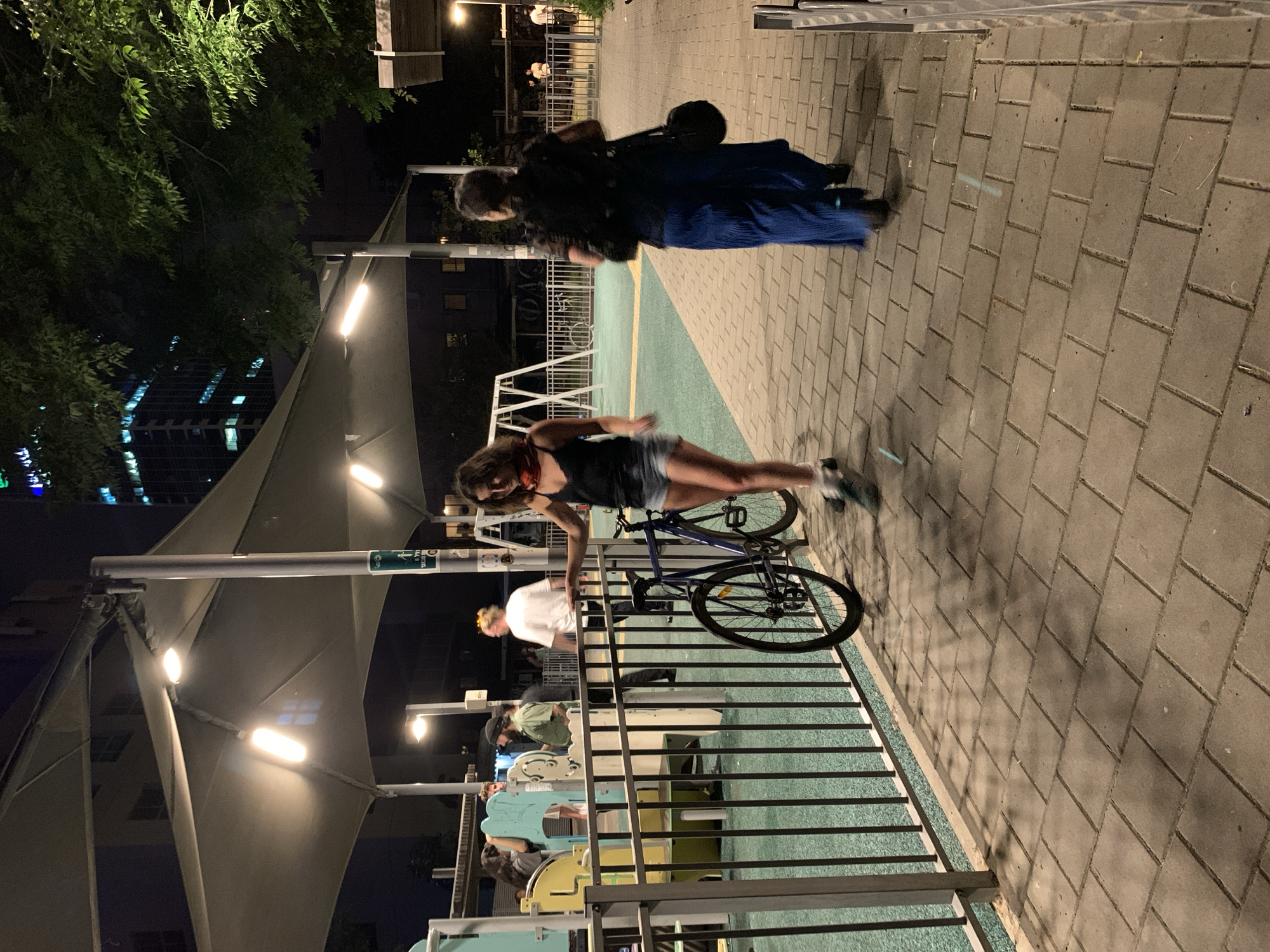 A night scene in what appears to be a park or recreational area of Tel-Aviv, Israel. The environment includes modern lighting fixtures, a shaded playground or activity area covered by a large fabric canopy, and surrounding trees. The ground is divided into a tiled walkway and a blue-green play surface.
There are several people in the image. In the background, two figures walk down the walkway. Closer to the foreground, a woman stands over a blue bicycle, appearing to lock it. Several other individuals are gathered at the activity area. The scene shows a casual, public space used for leisure activities during the evening.