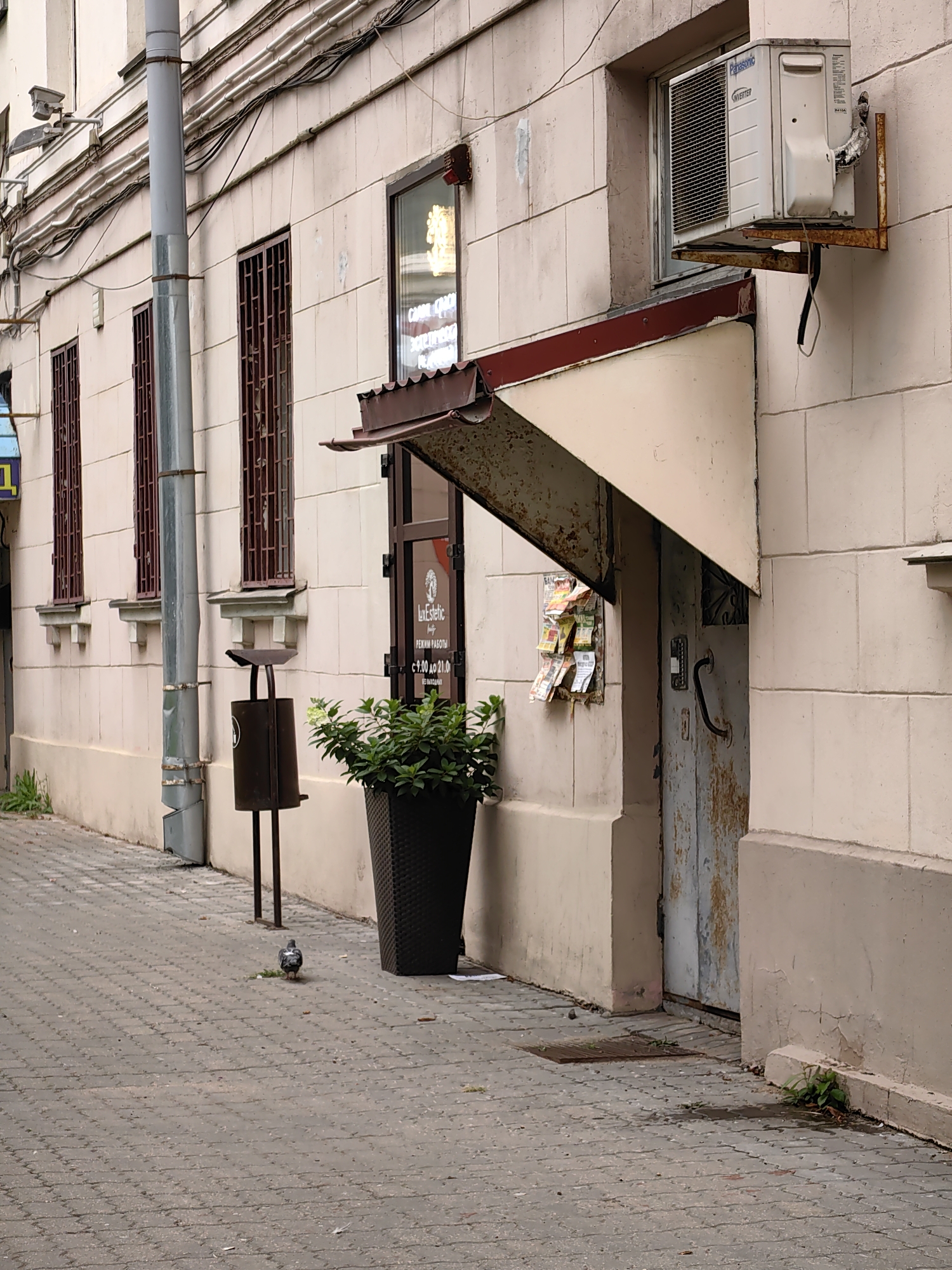 A pigeon walks on a paved sidewalk in front of a light-beige, tiled building in Minsk, Belarus. The sidewalk is made of interlocking grey pavers. To the left of the pigeon is a dark, cylindrical trash can on a stand, and further left is a tall, grey pipe. The building has several windows with dark metal grates. An air conditioning unit is mounted outside a window on the upper right side of the building. A weathered, beige awning extends over a metal door, also on the right side of the building. Several colorful flyers are attached to the wall under the awning. A large, dark planter with green foliage sits next to the awning and door.