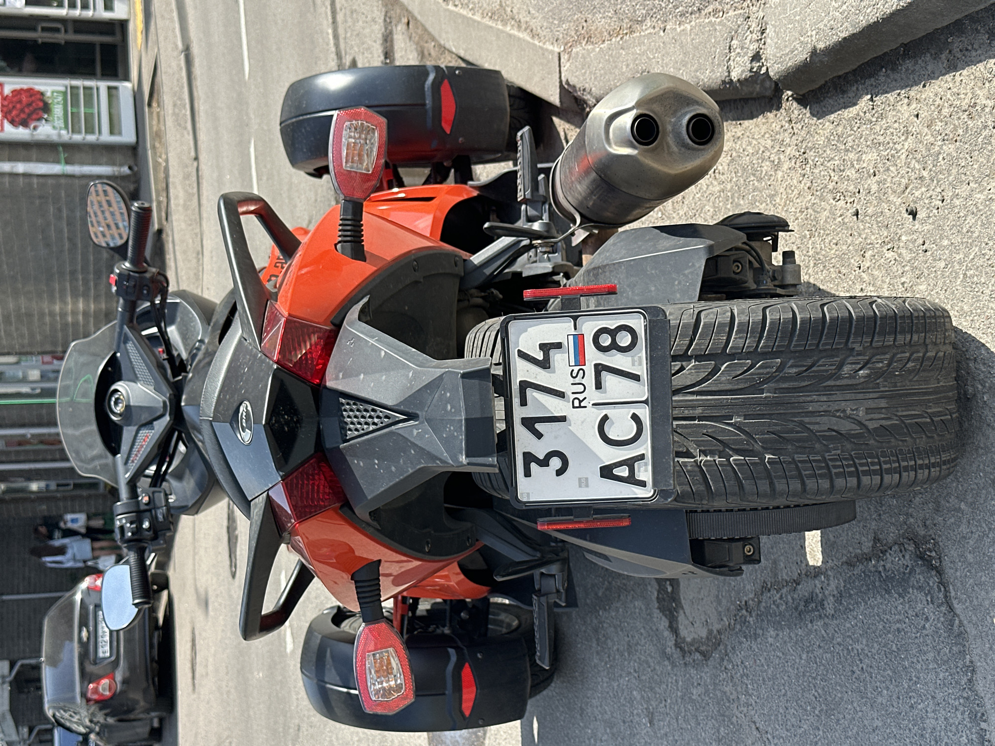 The image shows the rear of a three-wheeled vehicle, specifically a Can-Am Spyder, parked on a street. The license plate reads "3174 AC 78 RUS," indicating it is registered in Russia, specifically the 78 region (St. Petersburg and Leningrad Oblast). The vehicle has a distinctive dual exhaust pipe and a single rear tire. The vehicle is parked on a paved surface, likely a street or parking area, in Murino, Russia.
