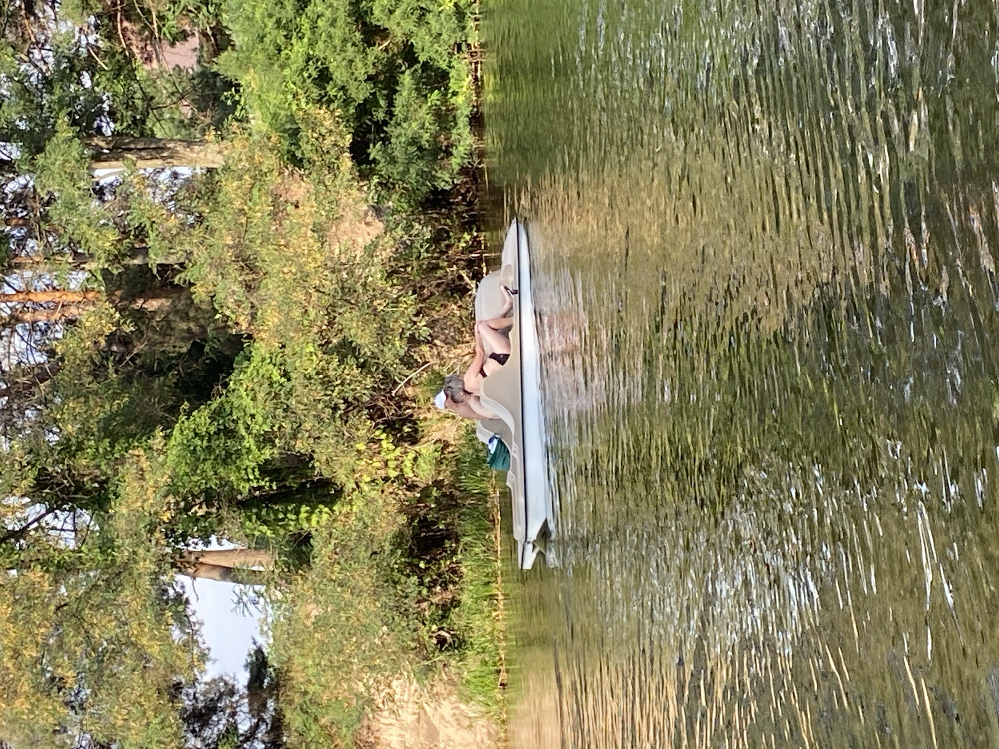 Two individuals are seen on a pedal boat in a body of water. The person in the foreground is wearing a black bikini and a white cap, reclining on the boat. The second person is partially visible behind them, also appearing to be reclining. The pedal boat is light beige and white and is positioned on the right side of the frame, moving away from the camera. The background consists of a lush green treeline and a sandy embankment on the left. The water's surface is rippled, reflecting the surrounding greenery and the sky. The scene appears to be a serene outdoor setting, likely a recreational area within or near the city of Petrivka, Ukraine.