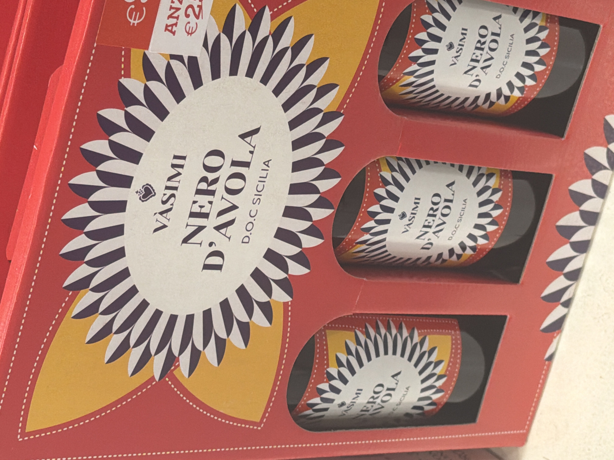 The image shows a red gift box containing three bottles of wine. The gift box has a decorative sunburst pattern in yellow and black and white. Each bottle also has a label with a similar sunburst design and the text "VASIMI NERO D'AVOLA D.O.C SICILIA". A small orange tag with text in a foreign currency is visible on the right side of the box. The boxes and bottles appear to be new and unopened. There is no indication of the location in the city of Ferrara, Italy.