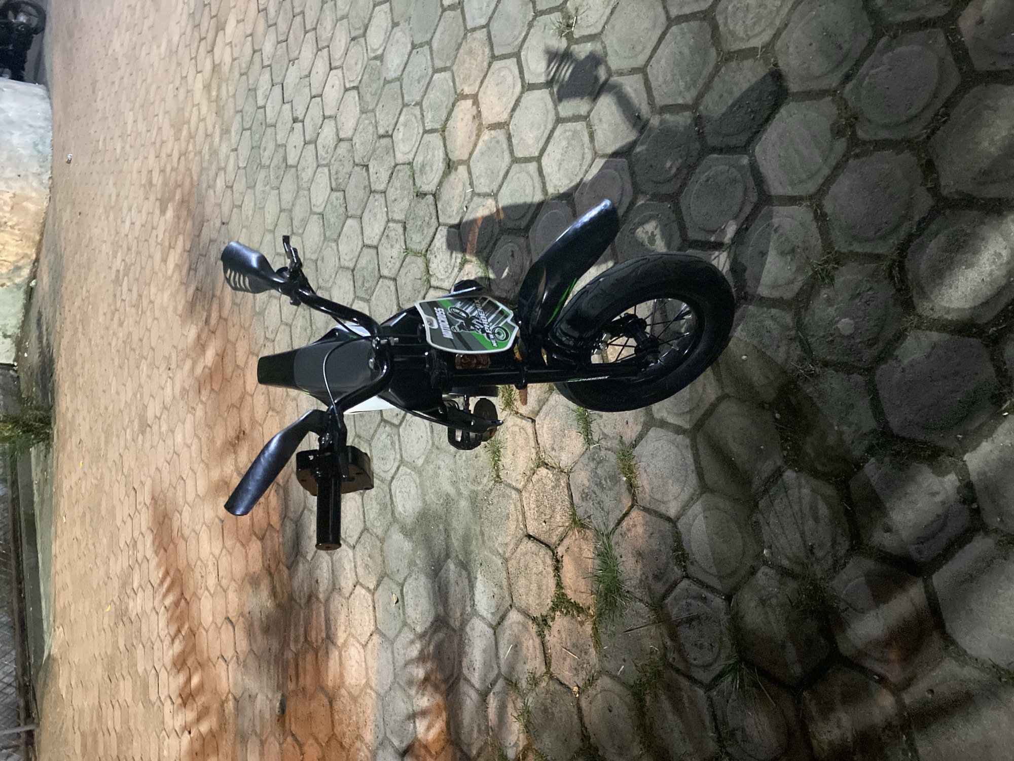 A black mini dirt bike is parked on a paved hexagonal stone surface at night in Lambaro Kueh, Indonesia. The bike is facing forward, with its handlebars turned slightly to the left. The front wheel is visible, as is the front fender. A green and white "MOTOCROSS NEW PHOENIX" sticker is affixed to the front of the bike. The pavement is illuminated by artificial light, casting shadows from the bike and surrounding objects. In the background, a light-colored wall and a fence are visible. Some sparse vegetation can be seen growing between the paving stones.