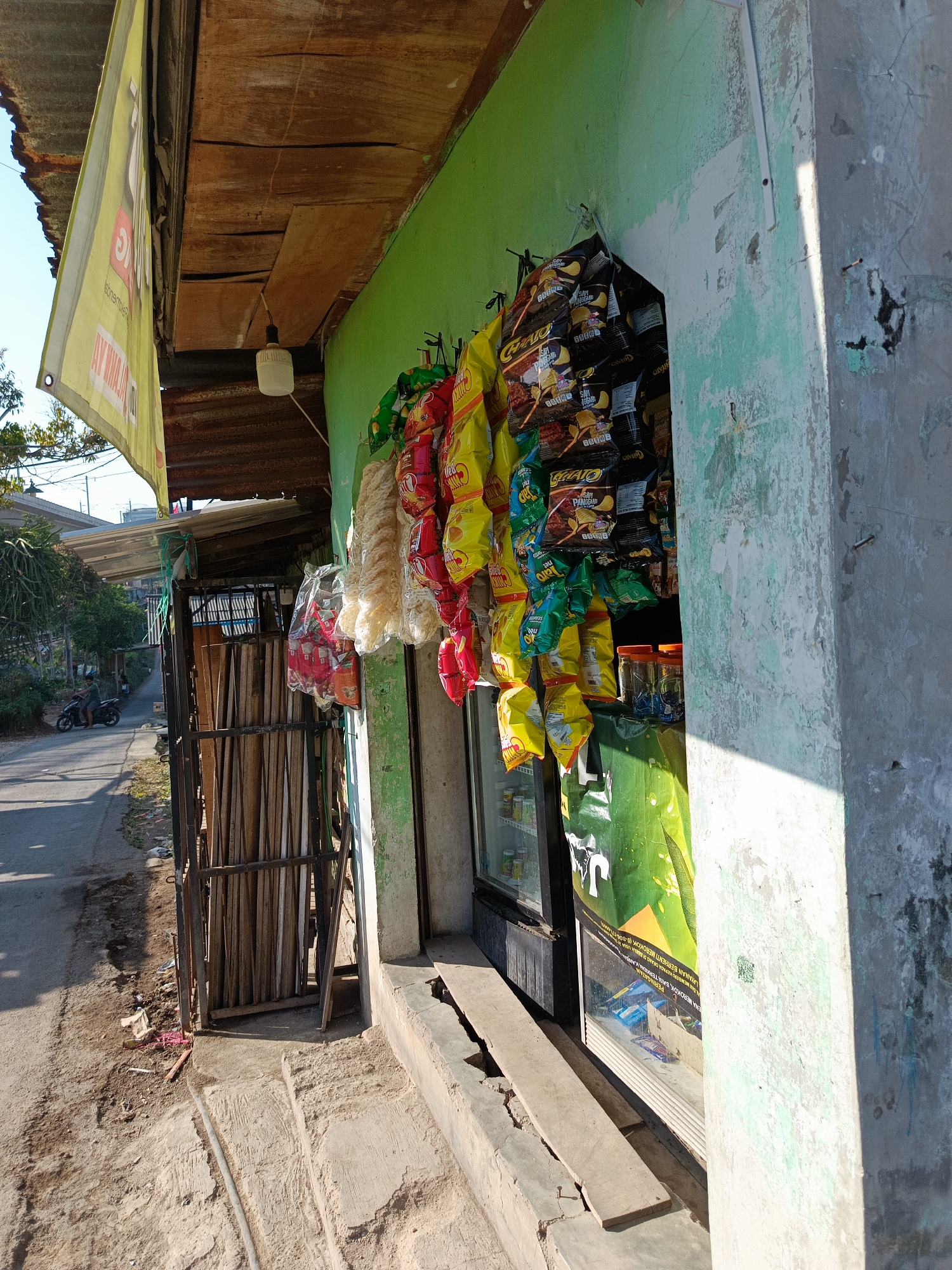 A street-side convenience store in Margajaya, Indonesia, features a green wall adorned with hanging snacks. To the left, a yellow banner with text is visible. A metal gate holding wooden planks stands next to the store. The street in front is paved, with a person on a motorcycle passing by. The scene is illuminated by natural sunlight.