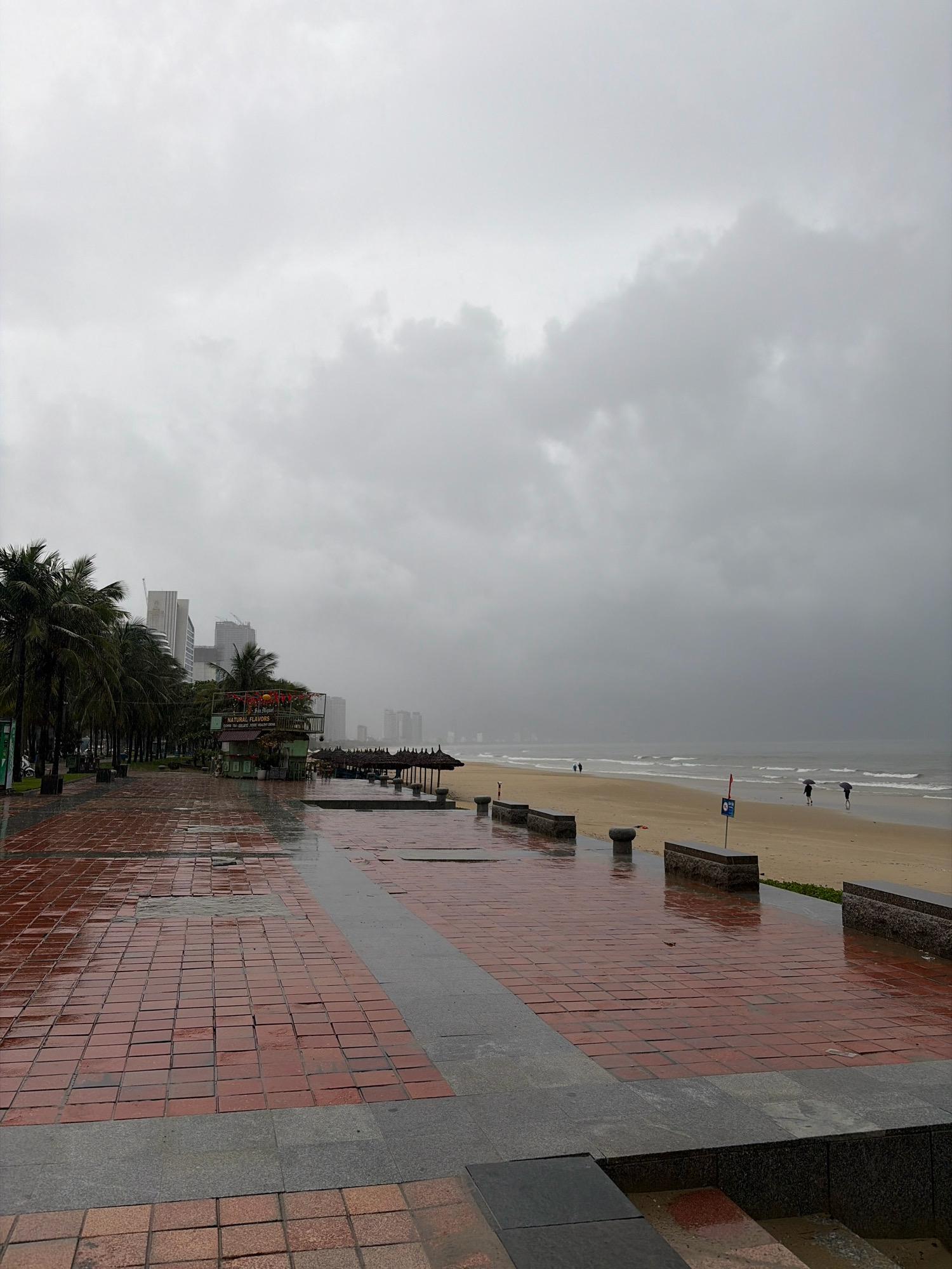 The image depicts a cloudy and overcast day at a beach in Da Nang, Vietnam. In the foreground, a wet, red brick pathway with grey stone borders reflects the dull light. The pathway leads towards a sandy beach where the ocean waves gently lap the shore. Further inland, a line of palm trees separates the pathway from a row of buildings, including some modern high-rise structures and what appears to be a small food stall or shop. A few people can be seen on the beach, some holding umbrellas, suggesting it may be raining or has recently rained. The overall atmosphere is one of subdued weather, with the grey clouds dominating the sky.