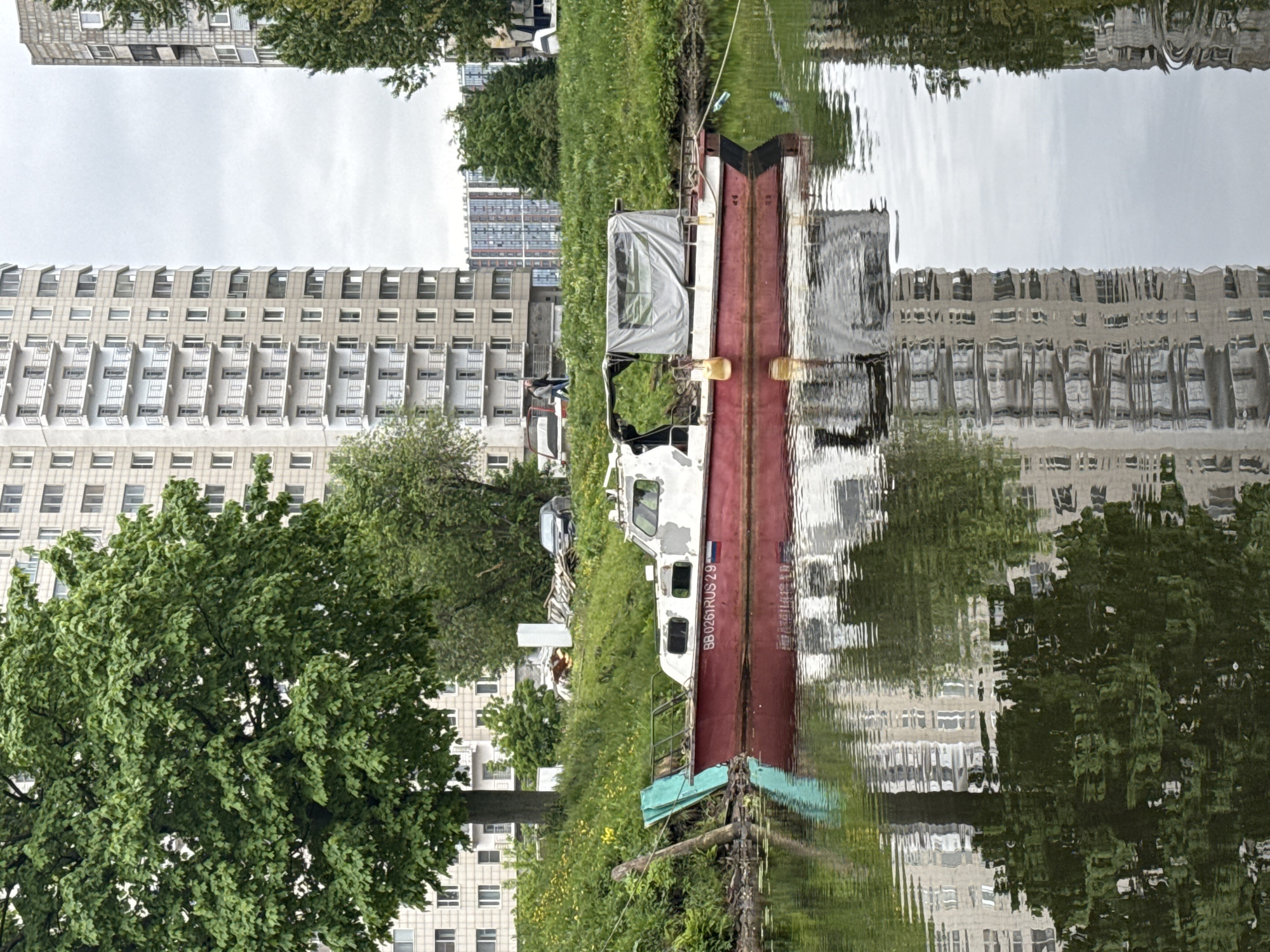 The image shows a narrow body of water in an urban setting, possibly a canal or small river. A dilapidated boat is moored along the grassy bank, with overgrown vegetation. The boat is white and red, with visible signs of wear and tear. The Russian flag is visible on the boat’s stern. Buildings, possibly residential apartments, are visible in the background, with a reflection of these buildings in the water. Based on the architecture and the Russian flag on the boat, the location is most likely Saint Petersburg, Russia. The image is rotated.