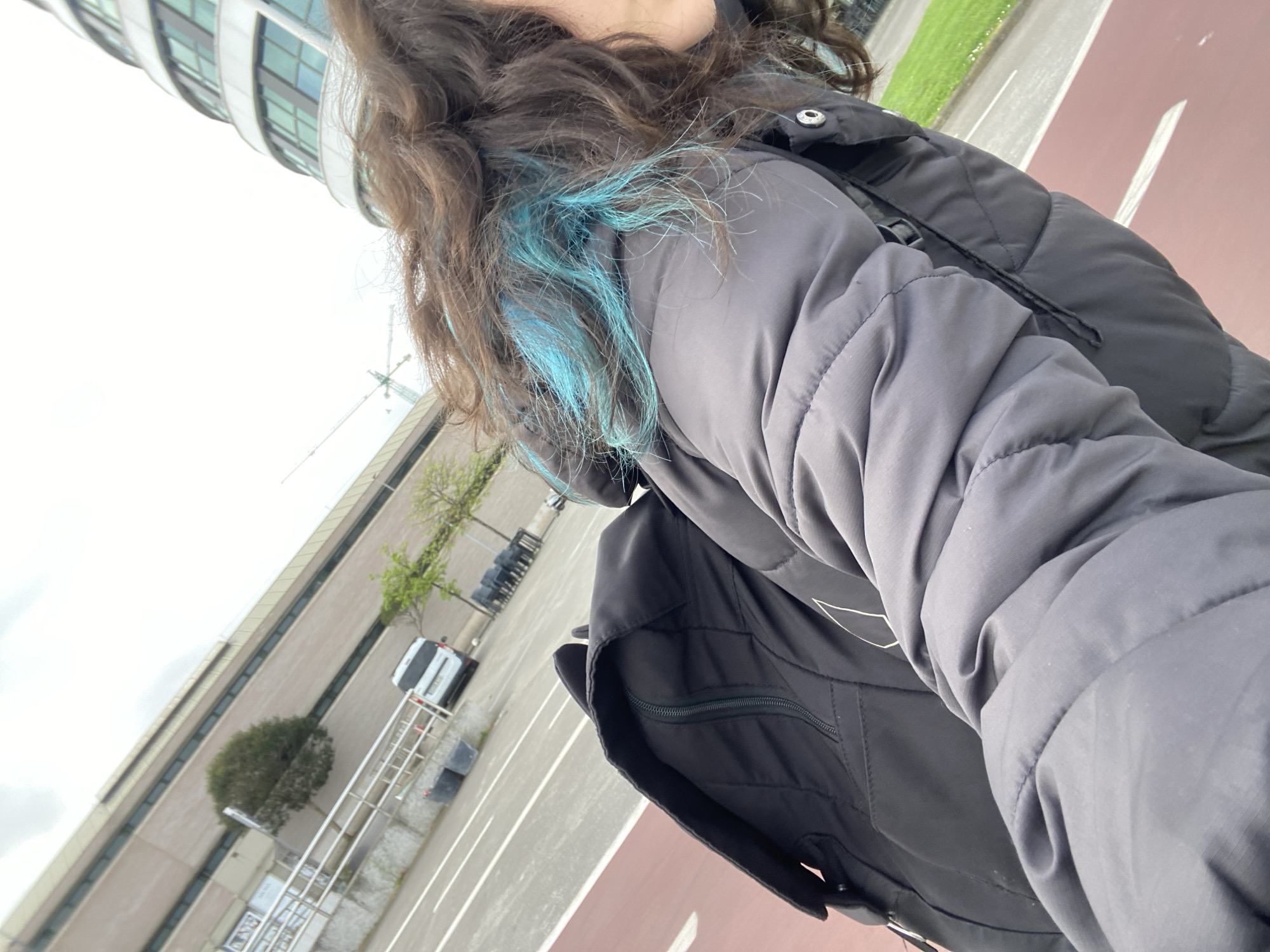 A first-person view shows a person wearing a gray quilted jacket with a black vest and has brown hair with blue streaks. The person is standing on a red asphalt path, part of a city street. In the background, a modern glass-facaded building rises, and a white car is parked along the side of the street. Some trees and bushes are visible, lining the sidewalk next to the building. A crane can be seen in the distance. The location is likely Gijón/Xixón, Spain.
