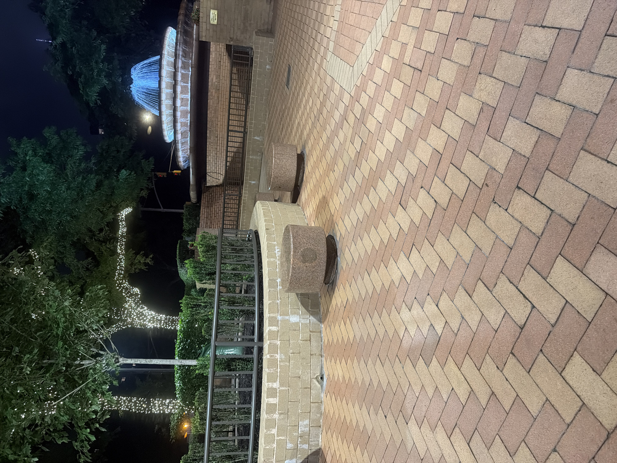 The image depicts an outdoor scene at night, featuring a tiered fountain with blue water illuminated. Surrounding the fountain are brick structures and lush green bushes. Two trees are visible, one with its trunk and branches wrapped in white string lights, and another with green foliage. The ground is paved with interlocking bricks laid in a herringbone pattern, primarily in shades of tan and reddish-brown. In the foreground, there are two cylindrical, granite-like planters. A dark metal railing separates the paved area from a raised planter bed with more greenery. The overall impression is of a well-maintained public space, possibly a park or plaza, illuminated for evening ambiance. The specific location within New York City cannot be determined from the provided image alone.