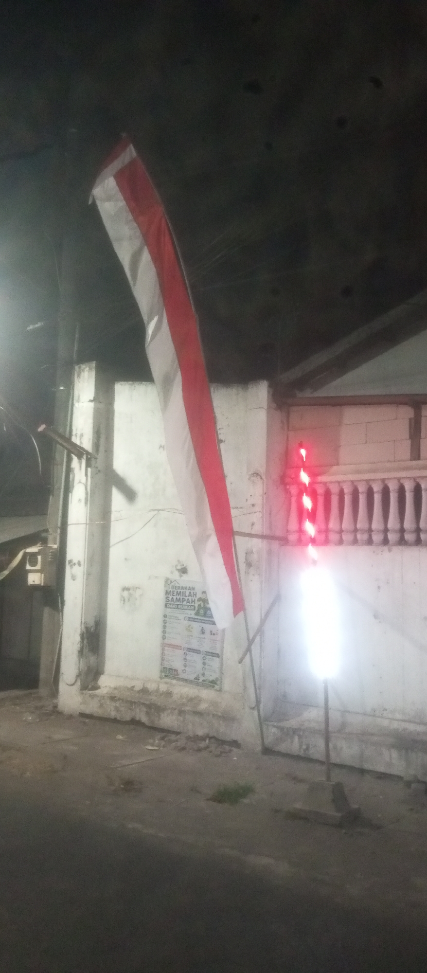 A red and white flag, likely the Indonesian flag, hangs from a pole on the left side of the frame. The flag is partially unfurled and illuminated by a bright white light on the right side of the frame, which also features a string of red lights above it. A white building with a textured, light-colored facade is visible behind the flag. The building has a decorative balustrade with vertical supports. A poster is affixed to the wall of the building, featuring text in Indonesian. The ground in front of the building appears to be asphalt or concrete, with some debris scattered on it. The scene is dimly lit, suggesting it is nighttime or a poorly lit area, potentially on a street in Kediri City, Indonesia, given the context. The overall impression is that of a street scene with patriotic or decorative elements.