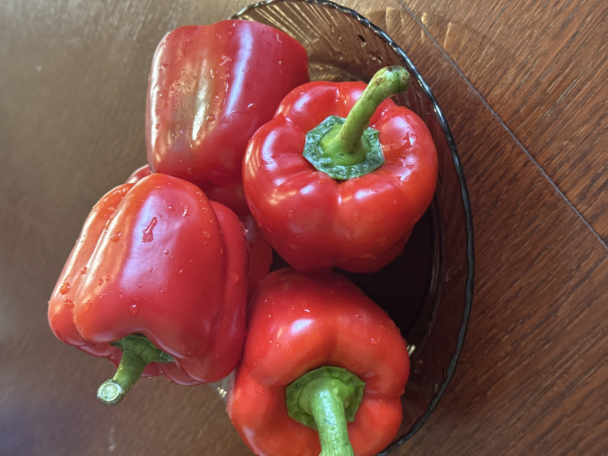 The media file is a top-down, close-up shot of a dark glass bowl filled with four bright red bell peppers. The peppers are wet, with visible water droplets on their surfaces, suggesting they have recently been washed. Each pepper has a vibrant green stem and is plump and glossy.
The bowl is placed on a wooden surface, likely a table, with visible wood grain running diagonally across the frame. The lighting appears to be natural, casting subtle highlights on the peppers and the bowl's rim. The overall scene suggests a simple preparation for cooking or a fresh display of produce. No people, text, or specific environmental cues beyond the wooden surface are discernible. The focus is entirely on the texture and color of the bell peppers within the bowl.