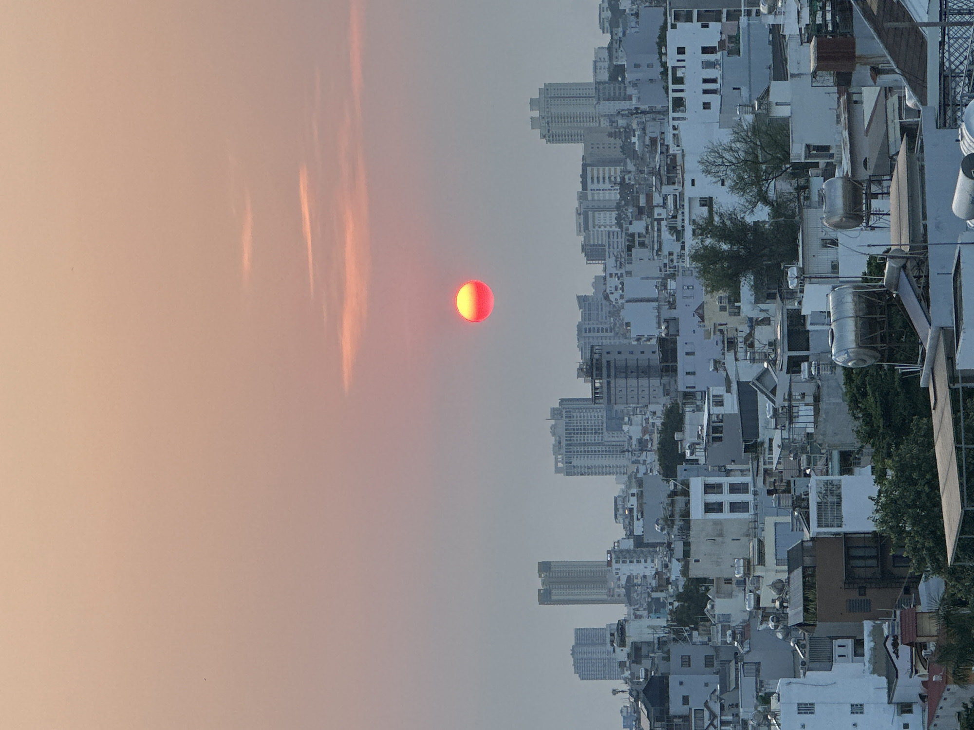 This image captures an urban landscape during either sunrise or sunset, centered on a striking red sun against a hazy sky. The scene depicts a densely populated city, identified as Ho Chi Minh City, Vietnam, with a multitude of buildings stretching across the horizon.
The sky occupies roughly two-thirds of the frame, transitioning from a soft, peachy-orange hue near the sun to a lighter, somewhat grayish-blue further away. Streaks of elongated, warm-toned clouds are visible, particularly near the sun, indicating atmospheric conditions like haze or light cloud cover. The sun itself is a prominent, vibrant red-orange orb, appearing slightly diffused due to the atmospheric conditions.
Below the expansive sky, the city's architecture is a complex tapestry of structures. Most buildings are light-colored, ranging from white to various shades of gray, with a mix of low-rise houses and taller, multi-story residential or commercial buildings. Many rooftops feature visible water tanks and other utility installations, characteristic of urban settings in the region. Scattered throughout the dense concrete jungle are pockets of green foliage and trees, providing a touch of nature amidst the urban sprawl. The overall impression is one of a bustling city at a tranquil hour, bathed in the soft, warm light of the setting or rising sun. No text is visible in the image.