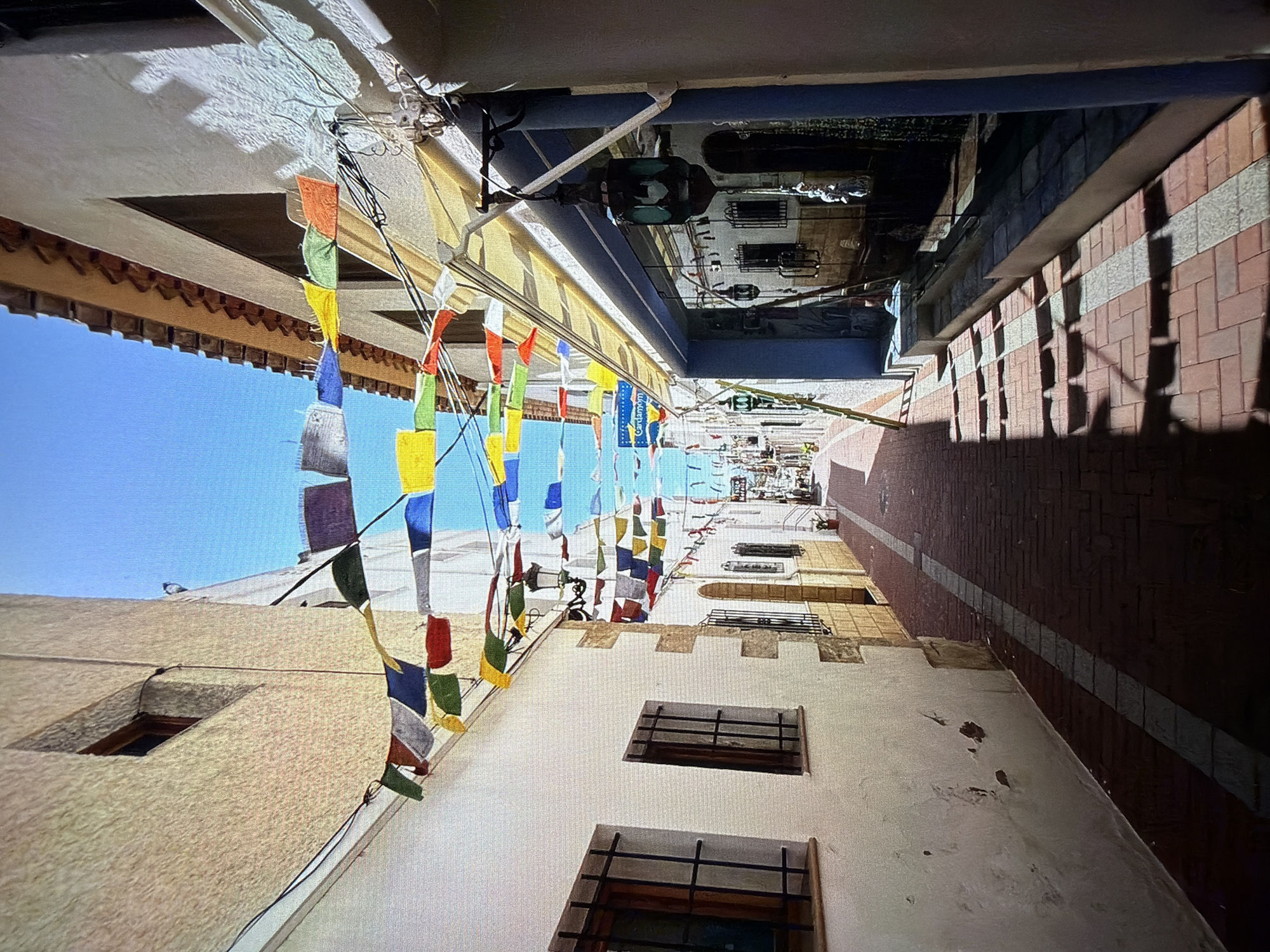 The original image, presented rotated 90 degrees clockwise, captures a narrow, brightly lit alleyway or street viewed from below, looking upwards. The clear blue sky occupies the left side of the frame, while the ground, paved with reddish-brown bricks and interspersed lighter stone stripes, is visible on the right.
Numerous strings of colorful, rectangular flags, reminiscent of prayer flags, are strung horizontally across the alley. These flags display a spectrum of colors including red, orange, green, yellow, blue, white, and grey, creating a vibrant and festive overhead canopy.
On the upper portion of the image (which corresponds to the buildings lining the left side of the alley when viewed correctly), white-washed structures with sections of terracotta roof tiles are prominent. A distinctive blue awning or overhang is visible further down the street. A decorative black street lamp with a glass casing hangs centrally above the alley. The lower portion of the image (representing buildings on the right side of the alley) also features white-washed walls with windows protected by black metal grates.
Strong, well-defined shadows cast by the buildings and flags across the ground indicate bright sunlight, suggesting the time of day is midday or early afternoon. Numerous electrical wires are visible crisscrossing between the buildings. A blue sign with white text, partially obscured, reads "Cardamom," suggesting a shop or eatery.
The scene depicts a charming, historic urban setting, characteristic of towns like Blagoevgrad, Bulgaria, as indicated by the context. No people are visible, and the overall impression is one of peaceful, sunny alleyways adorned with traditional decorations.