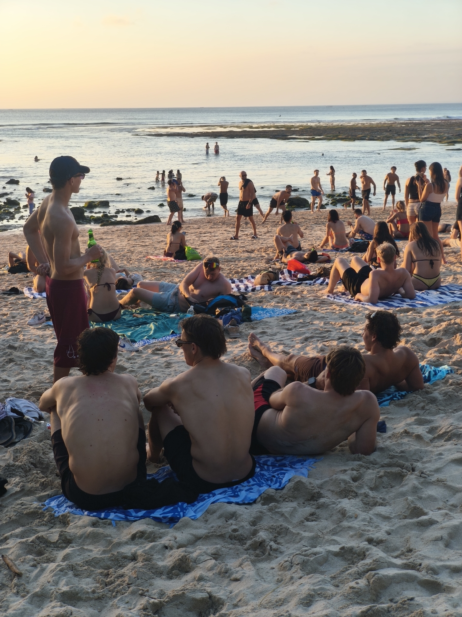 In Kuta Selatan, Indonesia, a group of people are gathered on a sandy beach during what appears to be late afternoon or early evening. The tide is out, revealing rocks and shallow water near the shore, with the ocean stretching towards the horizon under a soft, hazy sky. Many individuals are casually dressed in swimwear, engaging in various activities. Some are lying on towels on the sand, sunbathing, or relaxing. Others are standing or walking along the water's edge, and a few are in the shallow water. In the foreground, several men are seated on the sand, facing away from the camera, their backs to the viewer. A man in the mid-ground, wearing a black baseball cap and maroon shorts, stands holding a green bottle. The overall atmosphere suggests a leisurely beach scene.