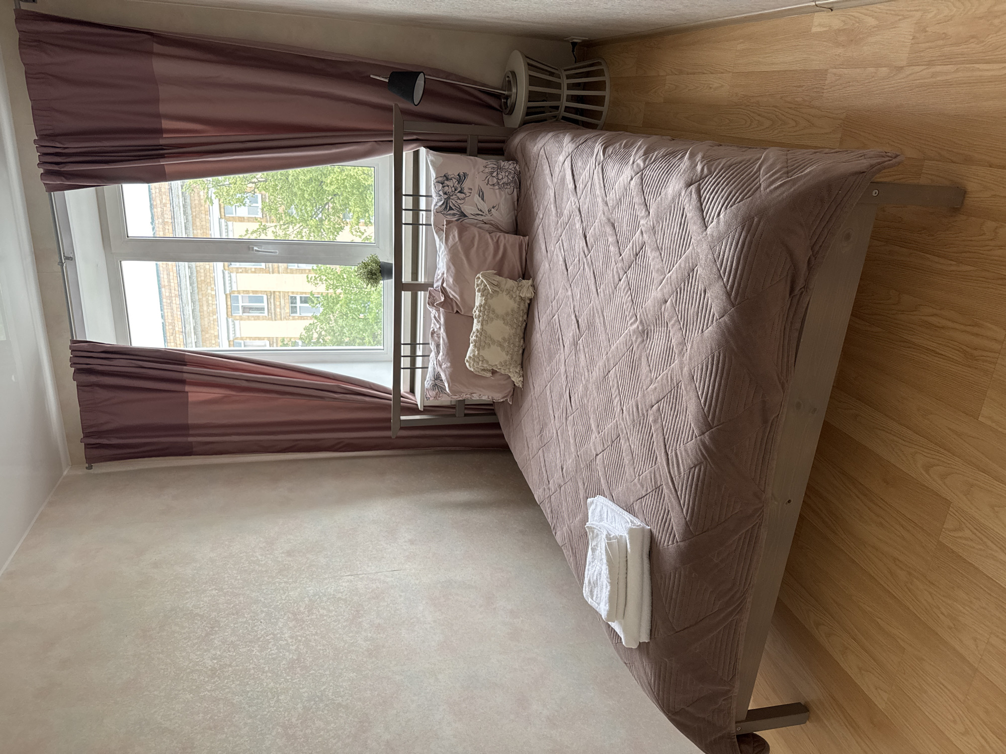 A bedroom in Veliky Novgorod, Russia features a queen-sized bed with a quilted mauve comforter and three pillows. One pillow has a cream-colored textured cover with tassels, another is mauve, and the third has a floral print. Stacked white towels rest at the foot of the bed. The bed frame is dark wood. A dark metal headboard extends upwards. To the right of the bed is a round, light gray side table with a black desk lamp on top. A window with mauve curtains and a white frame overlooks buildings and trees. The walls are a textured beige, and the floor is light wood laminate.
