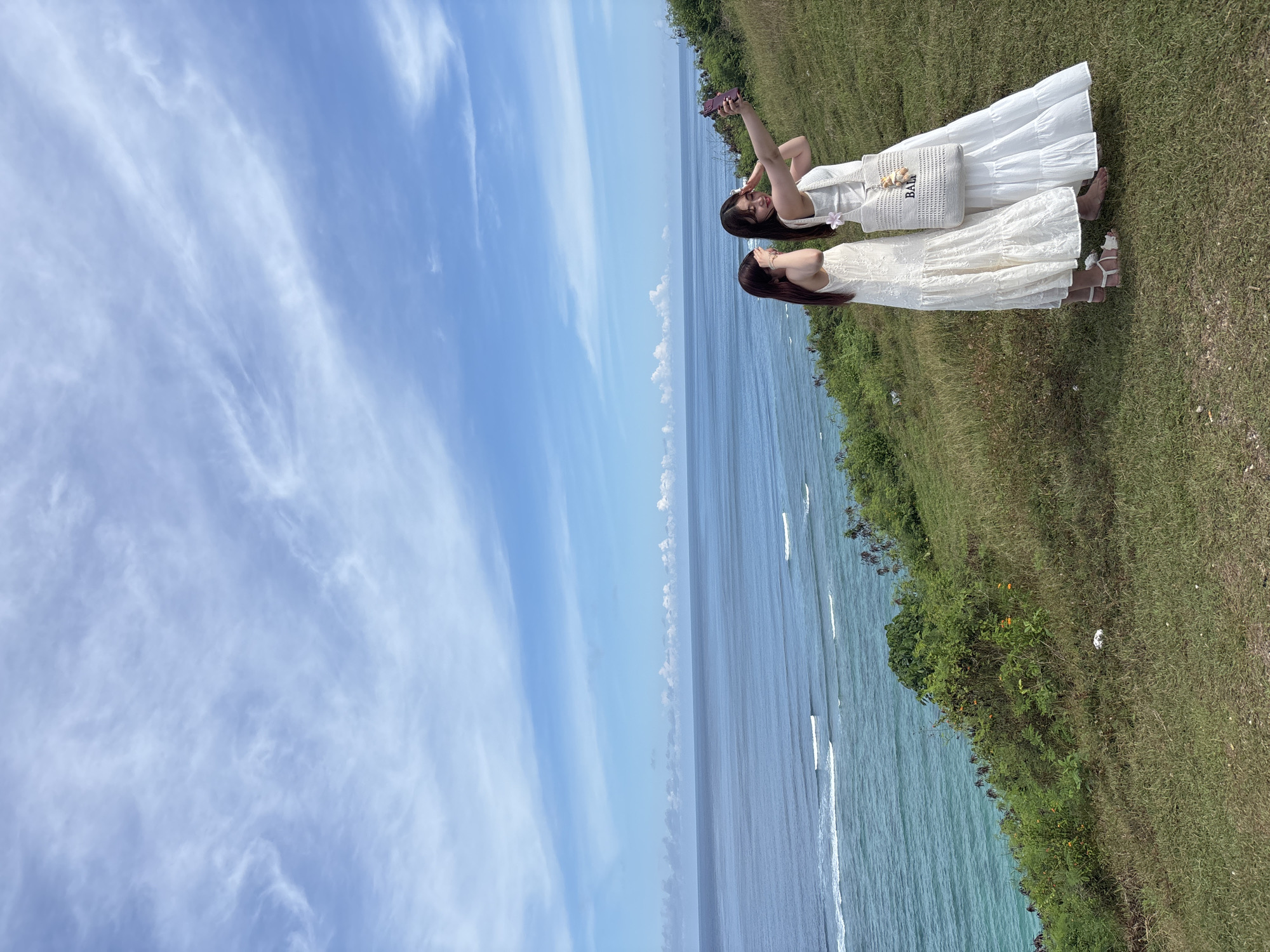 Two women stand on a grassy cliff overlooking the ocean under a blue, partially cloudy sky. The woman on the left is taking a selfie with her cell phone. Both women wear white dresses. The woman on the right carries a white "BALI" tote bag. The location is likely Kuta Selatan, Indonesia, given the coastal setting and bag.
