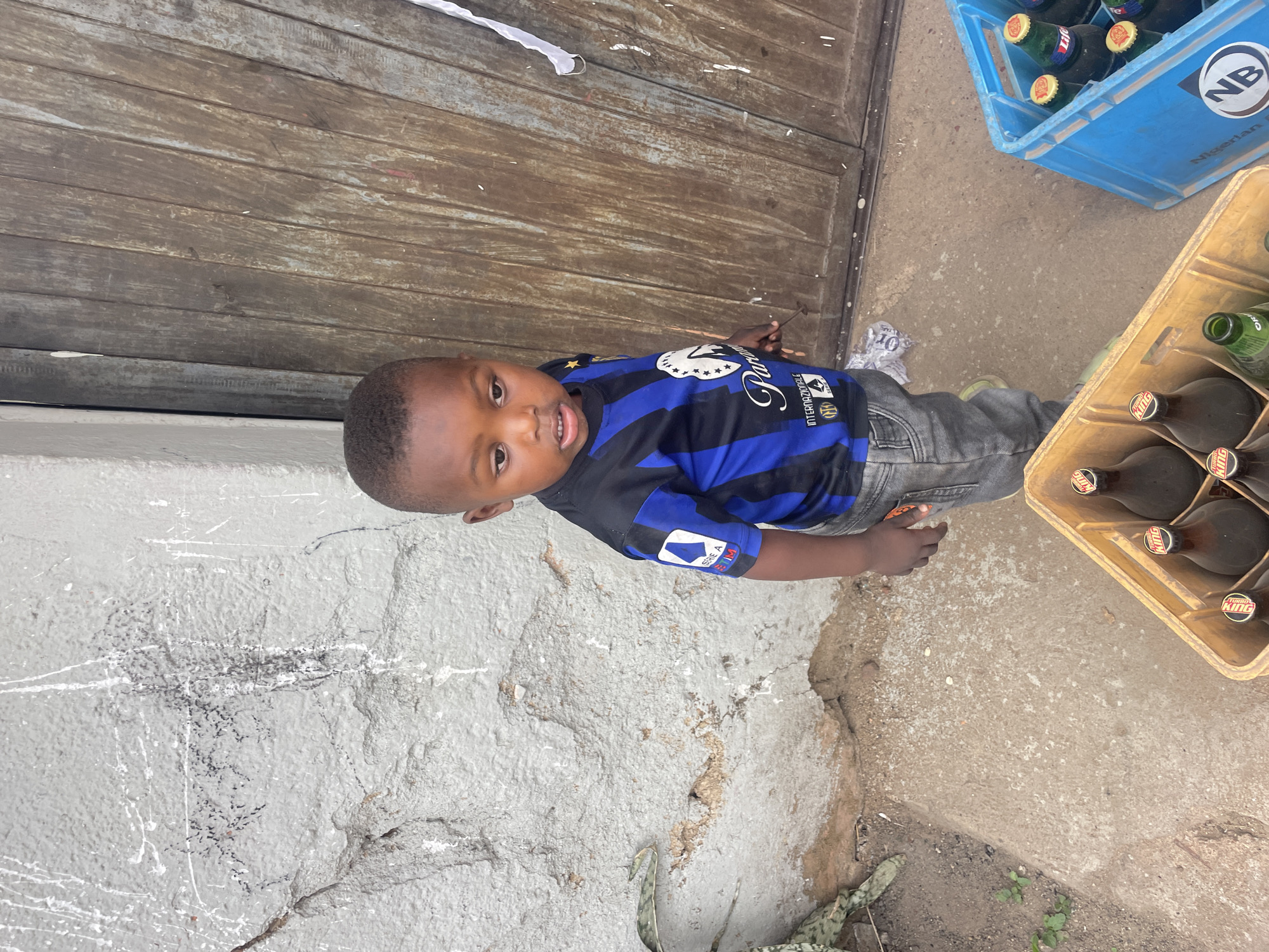 A young boy, appearing to be a toddler, stands outdoors, looking directly at the camera with a neutral and slightly curious expression. He is wearing a blue and black striped football jersey, identified as an Internazionale (Inter Milan) kit, with "Pirelli" partially visible on the front and "Serie A TIM" on a sleeve patch. He also wears grey jeans and light-colored shoes.
The setting appears to be an everyday street or yard scene in daylight. Behind the boy is a rustic, weathered wooden door or gate. To his right, two crates of bottled beverages are visible on the concrete ground. One is a blue plastic crate, partially filled with green bottles, some bearing "NB" for Nigerian Breweries and "Nigerian" text. The other is a light-colored plastic crate, holding dark bottles with red and yellow labels that clearly show "TURBO KING". To the boy's left, a light grey, textured wall or ground surface with some cracks and dark markings forms the foreground. The lighting suggests it is daytime. The scene depicts a common street-side or domestic environment typical of Jos, Nigeria.