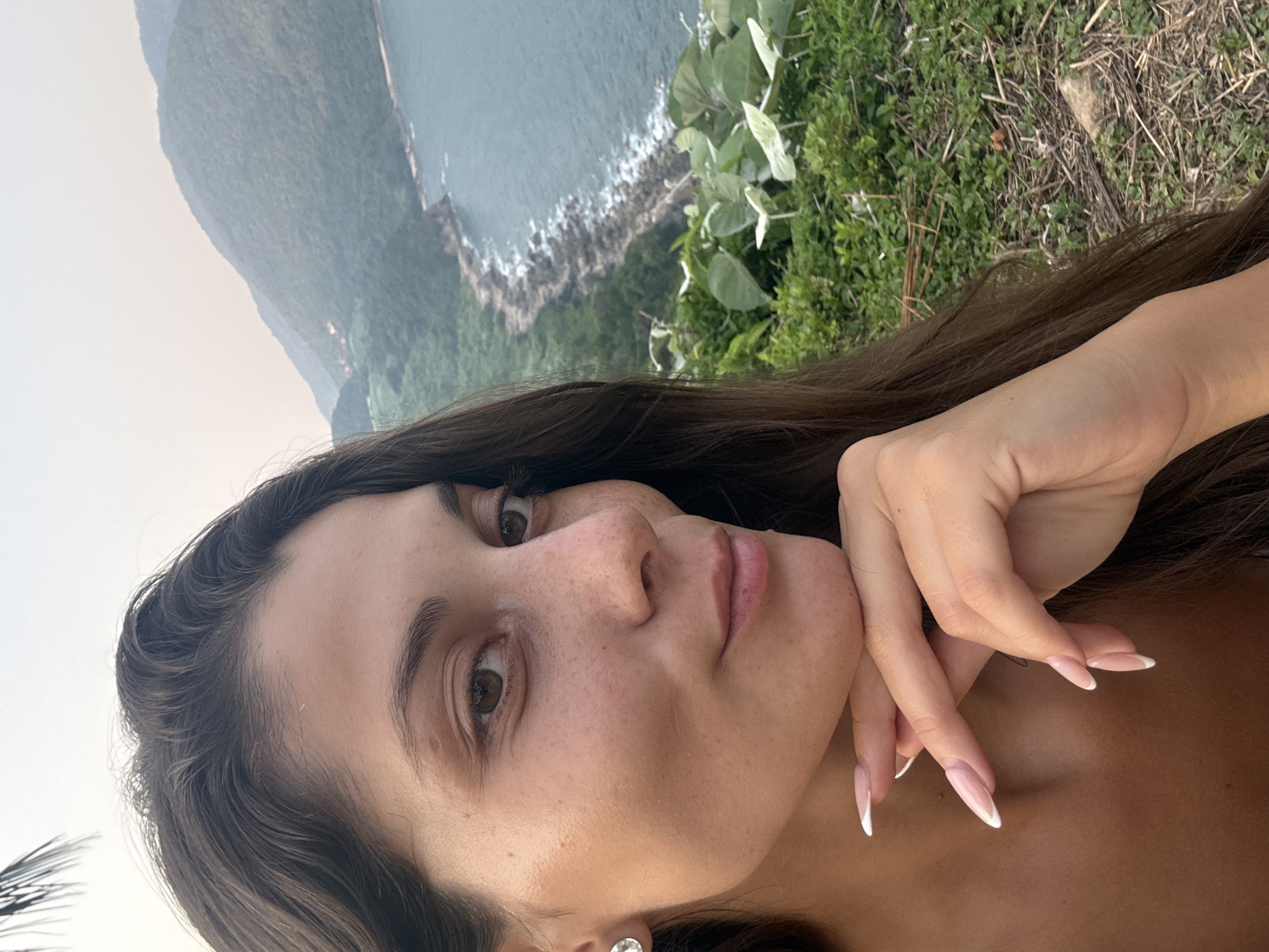 A woman poses for a selfie in Đà Nẵng, Vietnam. She has long, brown hair, light skin with freckles, and manicured nails. Her right hand is positioned near her chin. The background features a mountainous coastline, lush greenery, and a body of water.