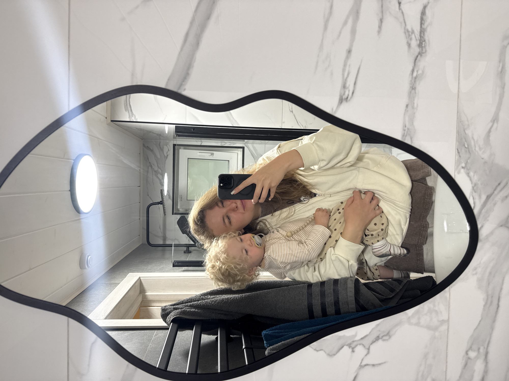 A woman is holding a baby and taking a selfie in a bathroom mirror. The woman has long blonde hair and is wearing a cream-colored hoodie and brown shorts. The baby has curly blonde hair and is wearing a striped onesie and polka dot pants. The bathroom has white marble walls and a black mirror. There is a towel rack with several towels on the left side of the image. In the background, there is a shower with a glass door and a small window. The location is the city of Prokudino, Russia.
