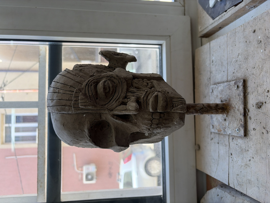 The image displays a forensic-style clay sculpture of a human head, bisected vertically, positioned indoors before a window overlooking an urban street in Sofia, Bulgaria.
The central subject is a dark beige clay bust, approximately human-head size, mounted on a rusted, dark brown metal rod with a square base, secured by four screws onto a light-colored wooden board. The sculpture's left half (viewer's left) depicts an anatomical skull, featuring an orbital socket, nasal cavity, and teeth. The right half (viewer's right) renders the muscular structure of a human face, including an eye with concentric lines, nasal cartilage, and half of a mouth with textured lips and exposed teeth. Horizontal and vertical score marks are visible on the forehead and temple areas of the anatomical half. The entire sculpture surface exhibits a rough, unfinished texture, consistent with raw clay.
The sculpture rests on a dusty wooden surface, likely a workbench or windowsill, with scattered specks of clay or debris. A portion of another wooden frame or object is partially visible on the right side of the wooden surface.
Through the clear pane of a white-framed window, an outdoor scene is visible. A light reddish-pink building facade occupies the background, featuring a white split-unit air conditioning compressor mounted on its wall. Below and to the left of the air conditioner, a light-colored sedan car (possibly silver or white) is partially visible, parked on what appears to be a street or parking area. Another building with a lighter, possibly beige, facade and a window is visible further back. The scene is illuminated by natural daylight.