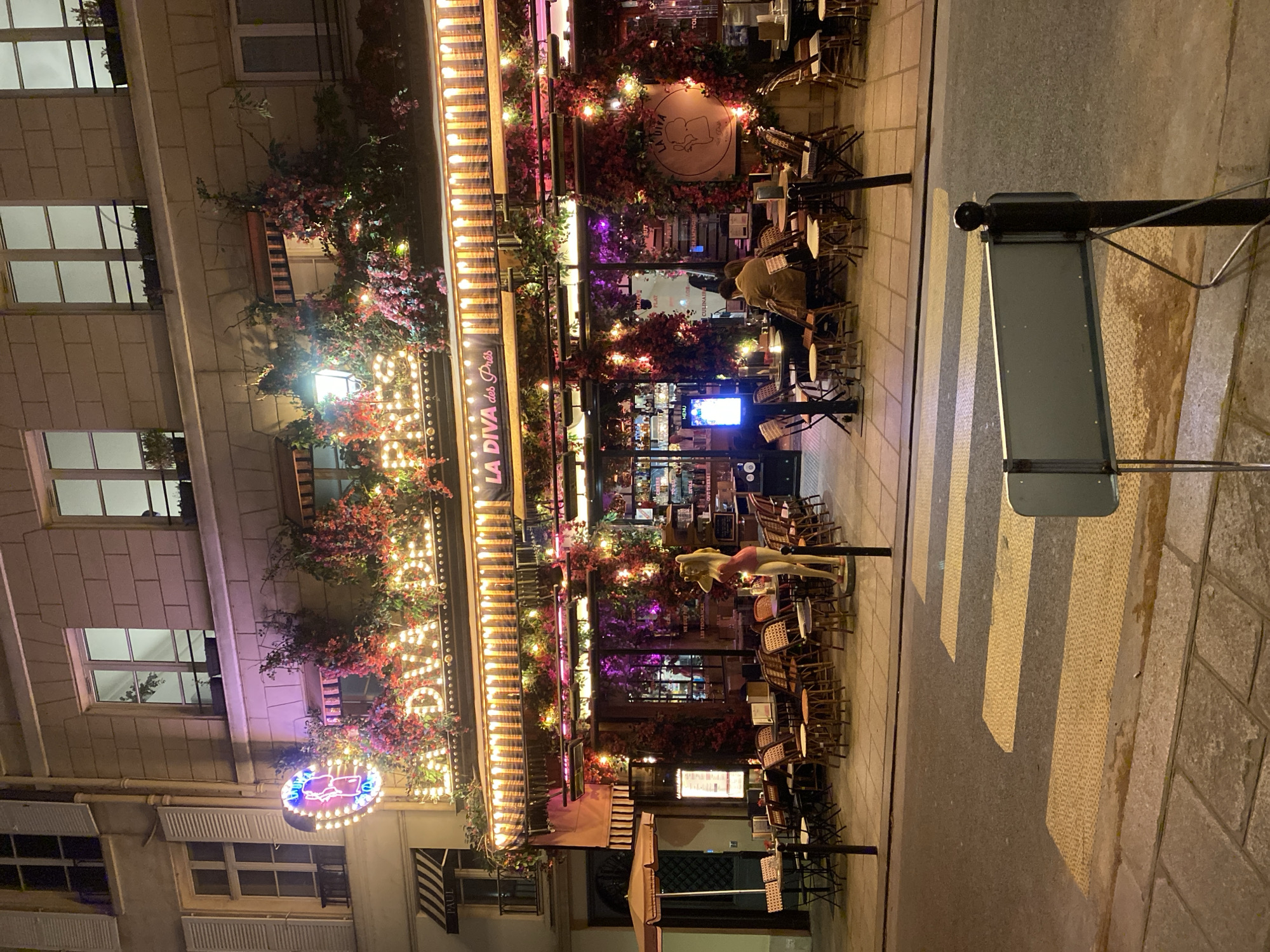 The image captures a vibrant nighttime street scene in Paris, France, featuring the exterior of a restaurant or cafe with extensive outdoor seating. The establishment, prominently named "LA DIVA des Près," is brightly illuminated with numerous string lights intertwined among decorative, bougainvillea-like plants that adorn its facade.
On the left side of the building, a large, colorful neon sign in the shape of a person is visible, adding to the festive atmosphere. A round sign above the entrance also displays "LA DIVA" along with a stylized caricature. Outdoor tables and chairs are arranged on the paved sidewalk, some appearing stacked, suggesting preparation for closing or a lull in business. One person, seen from behind in a light-colored coat, is seated at a table. Among the outdoor furniture, a standing mannequin or statue of a woman in a pink top adds a whimsical touch.
Further down the street, on the far right, a black stand holds a grey sign board, possibly displaying a menu or advertisement. The street features a clearly marked pedestrian crosswalk with yellow stripes. In the background, multi-story stone buildings characteristic of Parisian architecture, complete with windows and closed shutters, are subtly lit. A small striped awning with the text "PAUL" is visible on an adjacent building to the left, indicating another commercial establishment nearby. The overall ambiance is lively, welcoming, and romantic, typical of a European city evening. The visible text includes "LA DIVA des Près," "LA DIVA," "PAUL," and a small "MENU" sign visible through a window inside the restaurant.