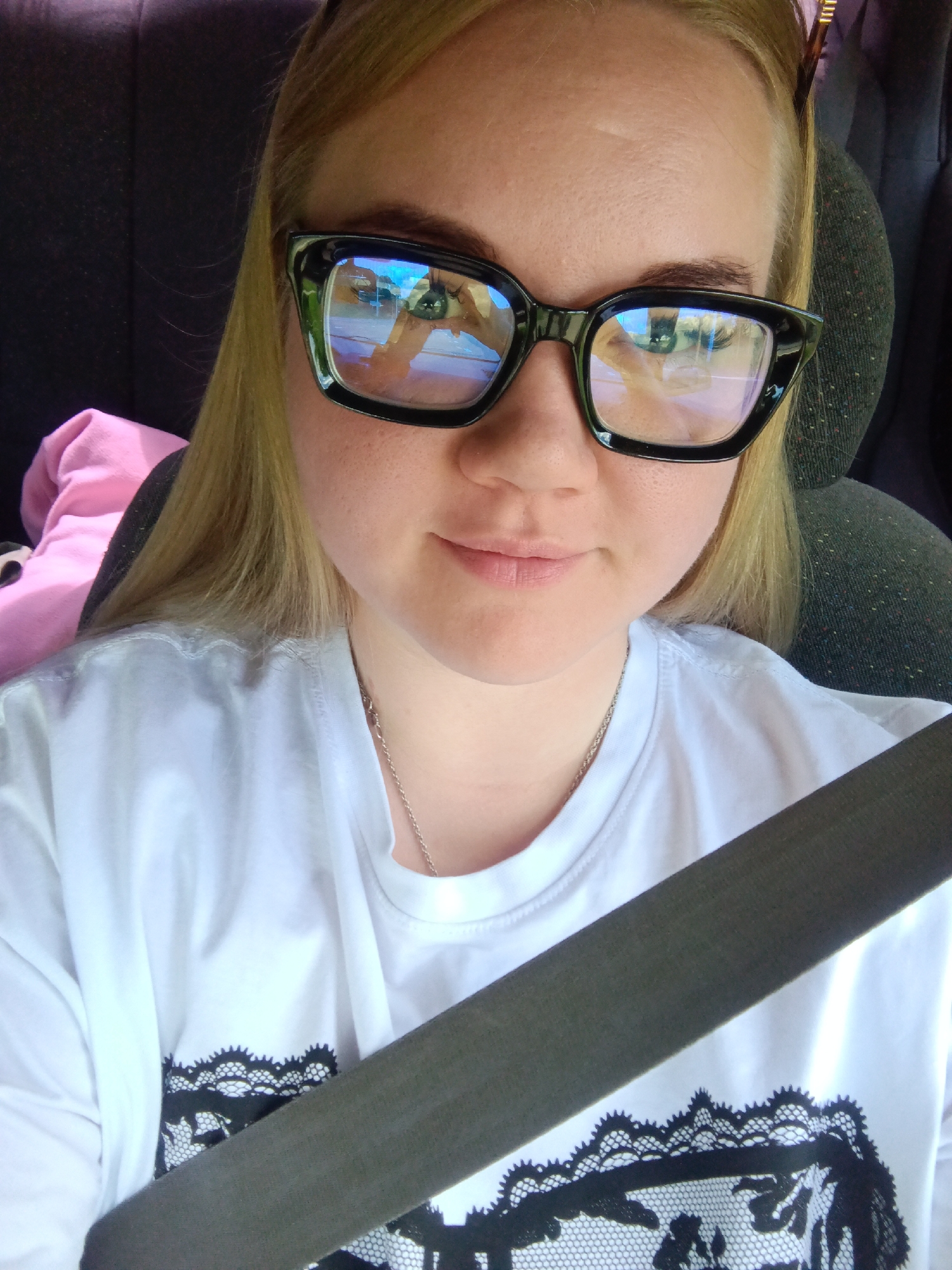 A young, fair-skinned woman with blonde hair is in the driver's seat of a car, wearing a white T-shirt with black lace detailing, and a silver necklace. She is wearing large black-framed glasses that reflect a bright outdoor scene. She is wearing a seatbelt. The location appears to be inside a vehicle, possibly in Votkinsk, Russia, based on the building structures and potential reflections in the glasses, though confirmation of the city requires further visual information.