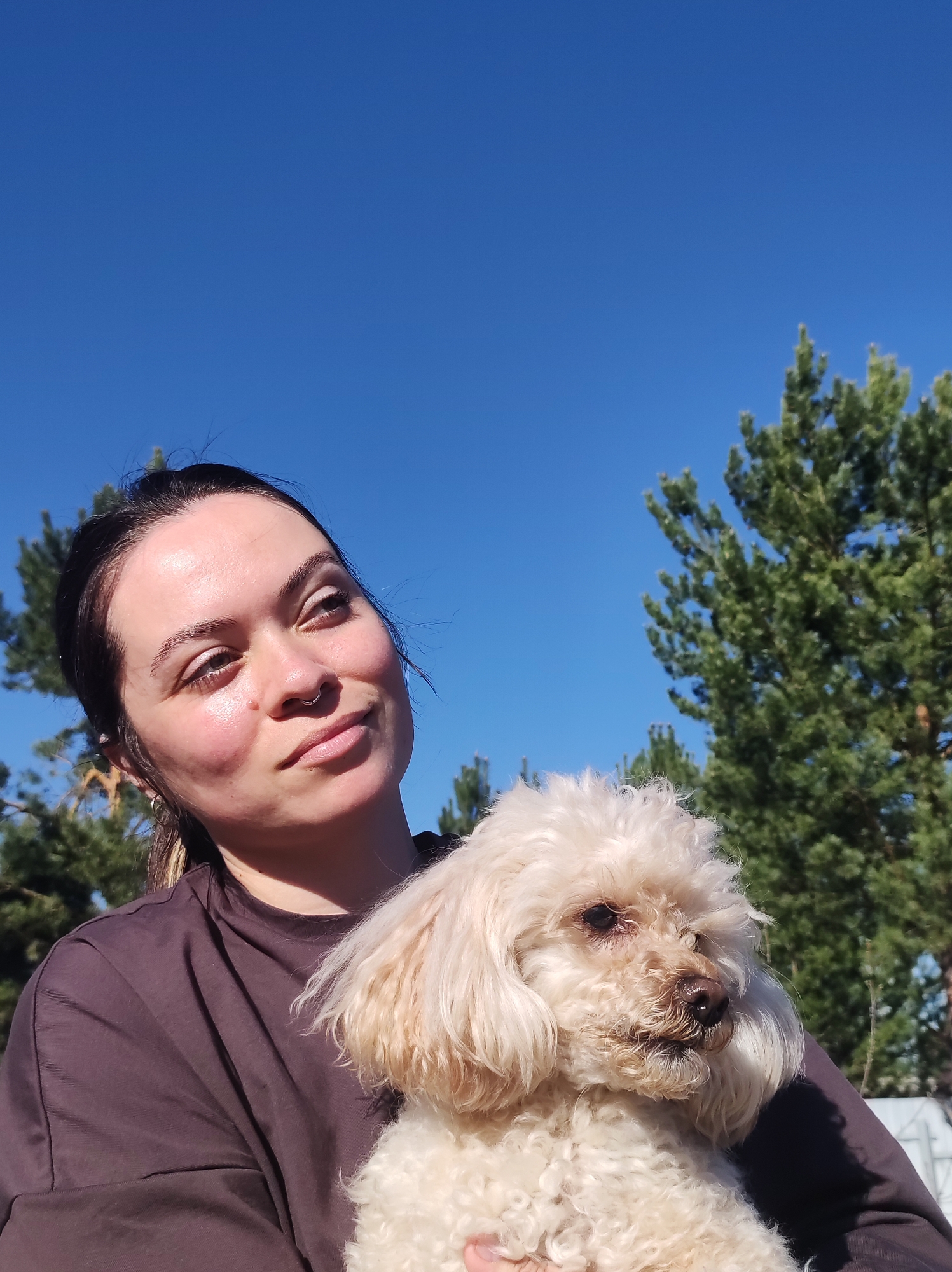 A woman is holding a light-colored poodle-type dog outside. The background features a clear blue sky and coniferous trees, likely in a park-like setting. The woman is wearing a dark t-shirt and has her hair pulled back. The location is indicated as Фоки, Russia. The woman is looking up with a slightly pensive expression.
