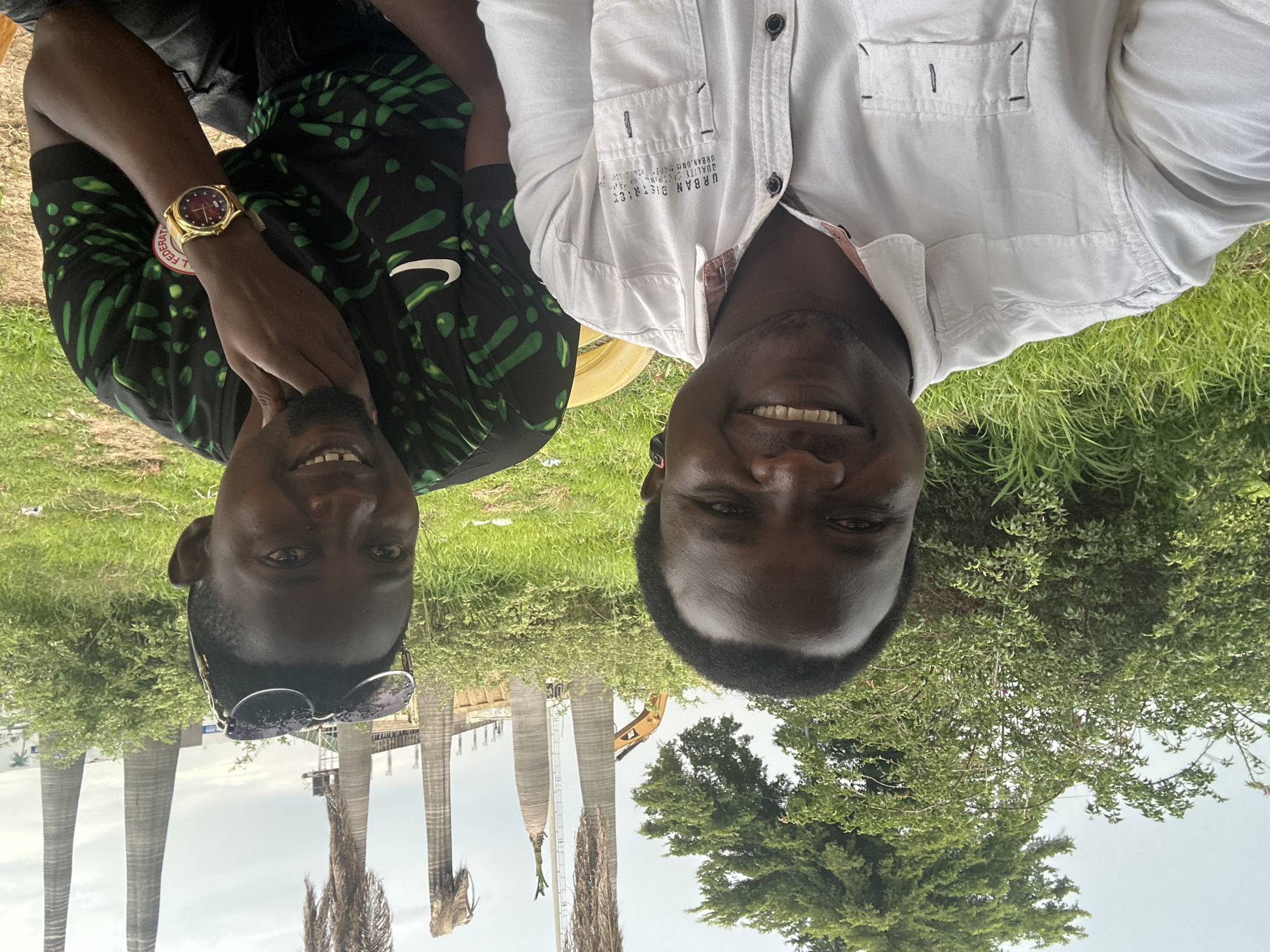 This media file captures a close-up, upside-down selfie of two men outdoors, with a blurry background of greenery and structures. The man on the left is wearing a black t-shirt with green patterns and a gold watch on his left wrist. The man on the right is wearing a white collared shirt with buttons and text on the pocket. Both men are smiling, appearing cheerful.
The setting appears to be a daytime scene with natural lighting, suggesting it could be a bright day, though the sky is overcast. The background shows lush green grass and trees, typical of an outdoor environment. In the distance, there are tall, cylindrical structures resembling silos or pillars, and a yellow construction vehicle is visible between them. These structures and the presence of construction equipment suggest an industrial or agricultural setting.
The text on the white shirt reads "URBAN DISTRICT" and "QUALITY THINKING". The gold watch has a circular face with Roman numerals and a red dial. The man on the left is also wearing sunglasses perched on his head. The overall mood is relaxed and friendly, as indicated by the subjects' smiles. The location context provided suggests this scene is in Dadi Riba, Nigeria.