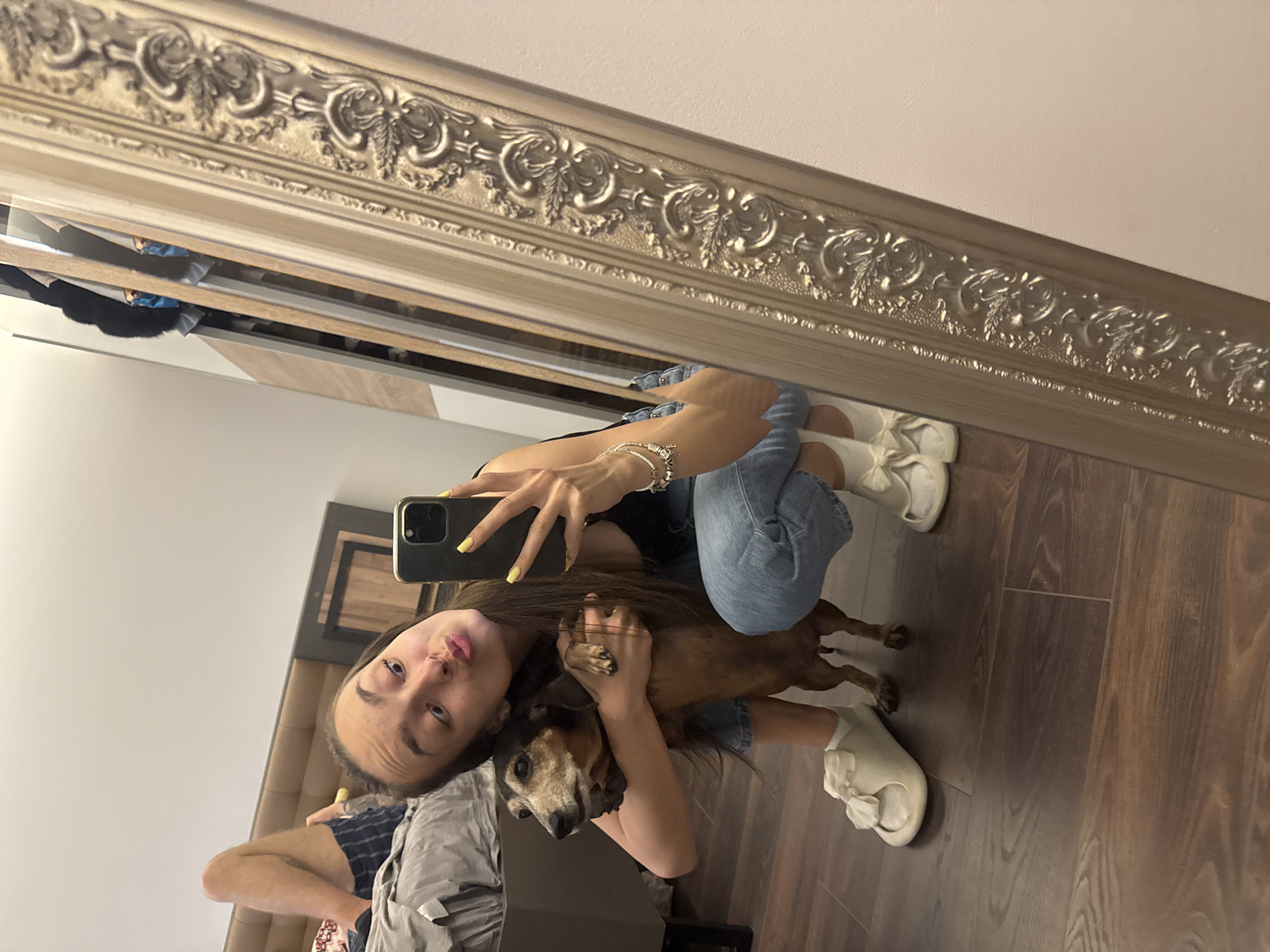 A young woman is taking a selfie in a room with wooden flooring. She is crouching and hugging a brown and black dachshund dog. She is wearing blue jeans, a black top, and white sneakers. The woman has long brown hair and is holding a black smartphone with yellow nail polish. The reflection in the ornate mirror above shows a glimpse of a bed and other furniture. Based on the style of the interior and the architectural details visible in the reflection, the location is likely a residential area in Sofia, Bulgaria.