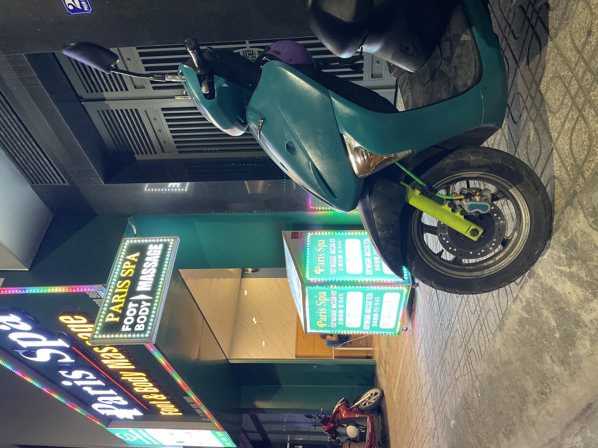 The image captures a vibrant nighttime street scene in Bắc Nha Trang, Vietnam, illuminated by various lights. In the foreground, a teal or turquoise-colored scooter is prominently parked on a textured concrete pavement. The scooter features distinct bright yellow-green front suspension components and a "HONDA" logo on its front panel. A purple helmet rests on its handlebars. Further down the street, a red scooter is also visible.
The scene centers around a business named "Paris Spa," advertised by multiple brightly lit signs. A large vertical sign displays "PARIS SPA" at the top, followed by "FOOT BODY MASSAGE" with arrows pointing to the services. Two additional cube-shaped signs provide details on "FOOT MASSAGE" and "BODY MASSAGE" services. Notably, these services are listed in several languages: English, Russian (МАССАЖ НОГ, МАССАЖ ТЕЛА), Chinese (足部按摩, 身体按摩), and Korean (발 마사지, 전신 마사지), indicating a diverse, international clientele.
The building behind the signs features a dark green facade with interior lighting visible through the entrance. A small sticker with the text "2 PHAM" is visible on an upper dark surface, potentially indicating an address or street name. The overall atmosphere suggests a lively urban area at night, common in tourist destinations like Nha Trang.