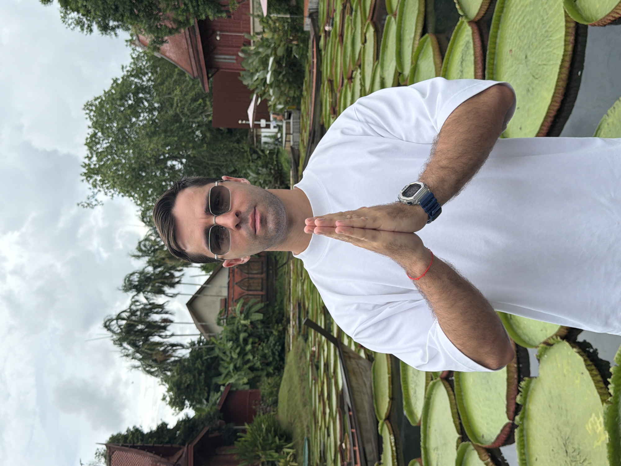 A man wearing sunglasses and a white t-shirt is photographed in a portrait orientation, positioned in front of a body of water filled with large, green lily pads. His hands are clasped in a prayer-like gesture at chest level. A silver wristwatch with a blue band adorns his left wrist. In the background, Thai-style buildings with red roofs and lush green trees are visible. The sky above is overcast, suggesting a cloudy day. The location is Thep Krasatti, Thailand.