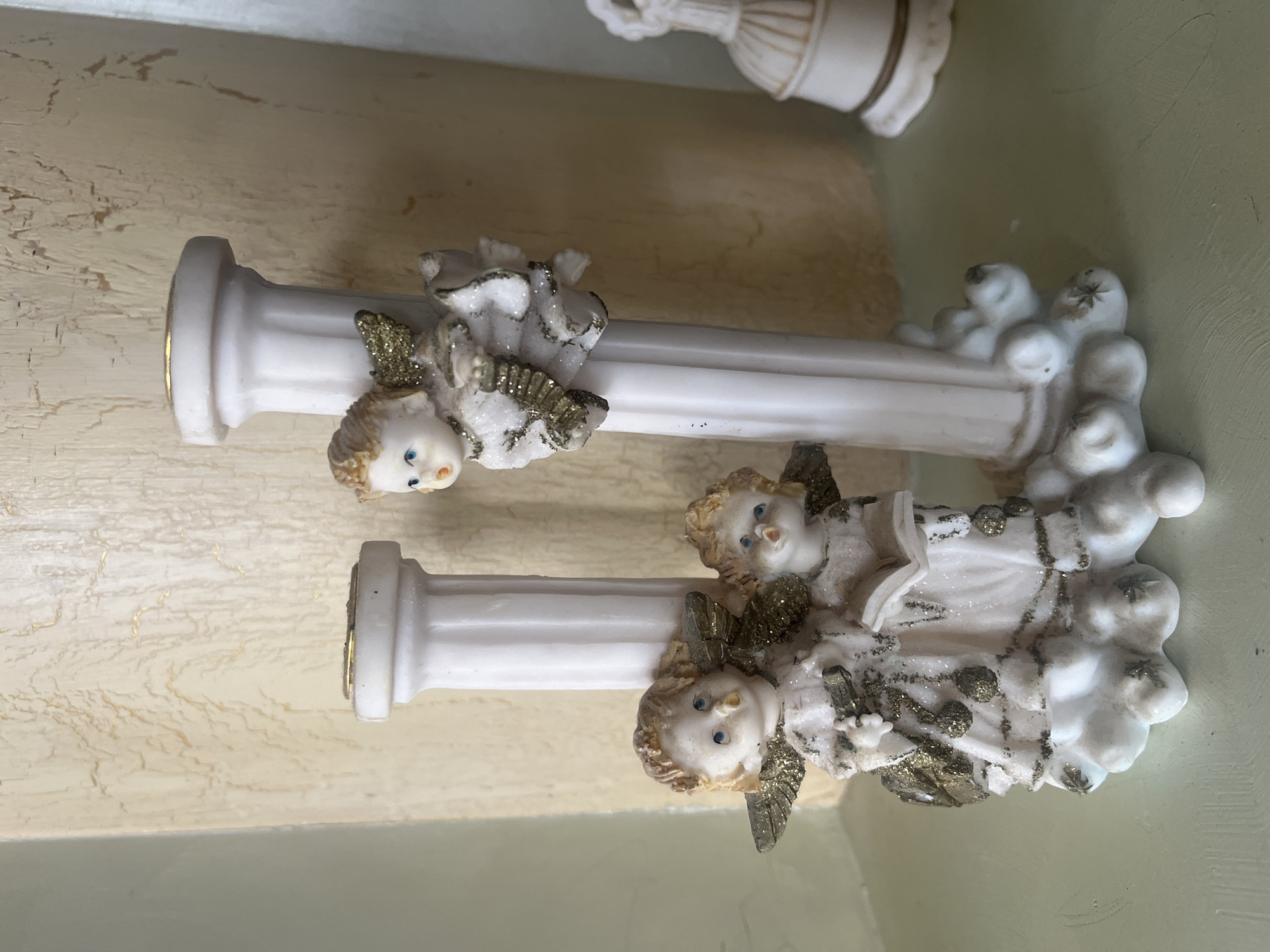 The image displays a decorative candlestick with three angel figurines. The candlestick is white and features two fluted columns. One angel is positioned on the left column, holding what appears to be a musical instrument, and another angel is to its right, reading a book. Both of these angels are adorned with gold glitter on their wings and clothing. A third angel is climbing on the right column, looking towards the left. All the angels have blonde hair and blue eyes. The base of the candlestick is decorated with clouds and gold accents. In the background, a textured, light-colored wall is visible. To the right, partially out of focus, is a white bell ornament. The candlestick is placed on a light green surface. The setting is an indoor environment, likely a shelf or mantelpiece. There is no discernible information in the image to place it in Tarnivtsi, Ukraine.