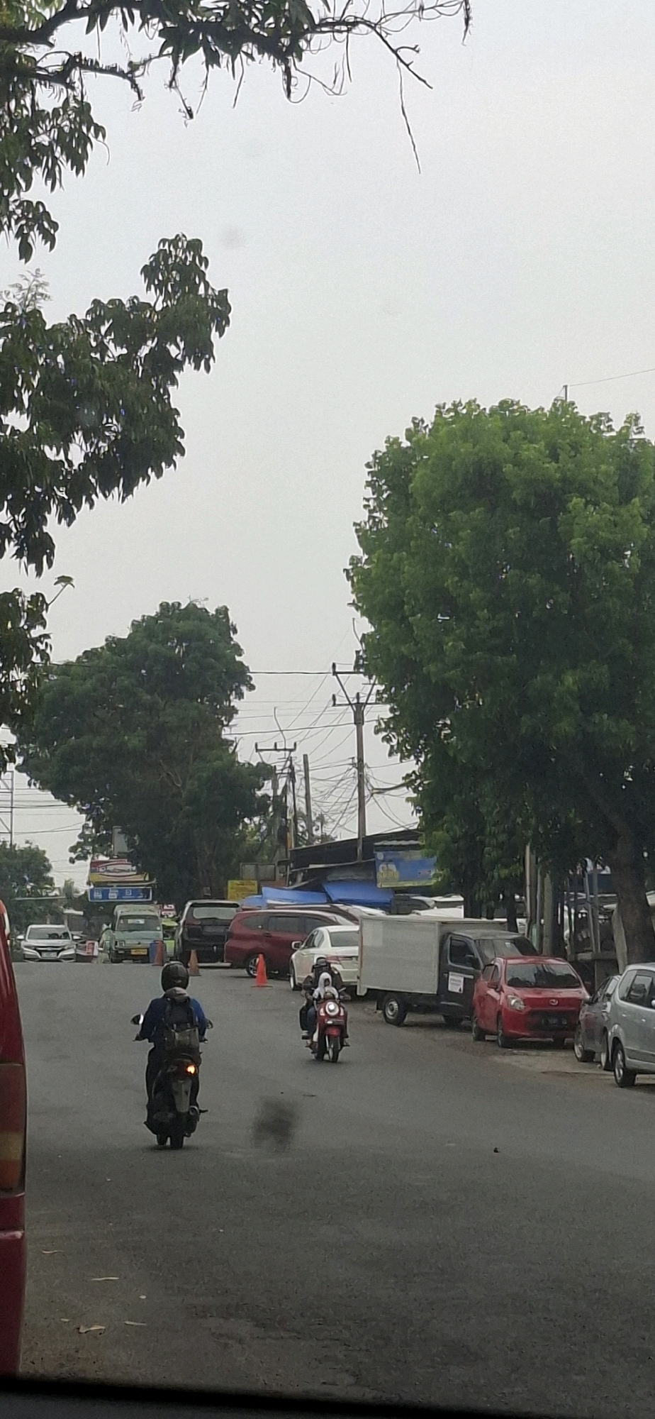 The image depicts a street scene in Rancagoong, Indonesia, characterized by moderate traffic and lush greenery. The sky is overcast, suggesting a cloudy day. In the foreground, the rear of a red vehicle is visible on the left. A motorcyclist wearing a blue jacket and a black backpack is riding away from the camera, with exhaust fumes emanating from the motorcycle. Further down the road, another motorcycle carries two passengers, one appearing to be a driver and the other a passenger, both wearing light-colored clothing and helmets.
The street is lined with various parked vehicles, including a red car, a white car, a silver car, and a white truck with a closed cargo box. Orange traffic cones are placed along the side of the road. In the background, trees with dense green foliage dominate the landscape, with some electrical poles and wires visible between them. Signs and structures that appear to be part of roadside businesses or residences are also present. The overall impression is a typical urban environment in Indonesia, where natural elements and human activity coexist.
