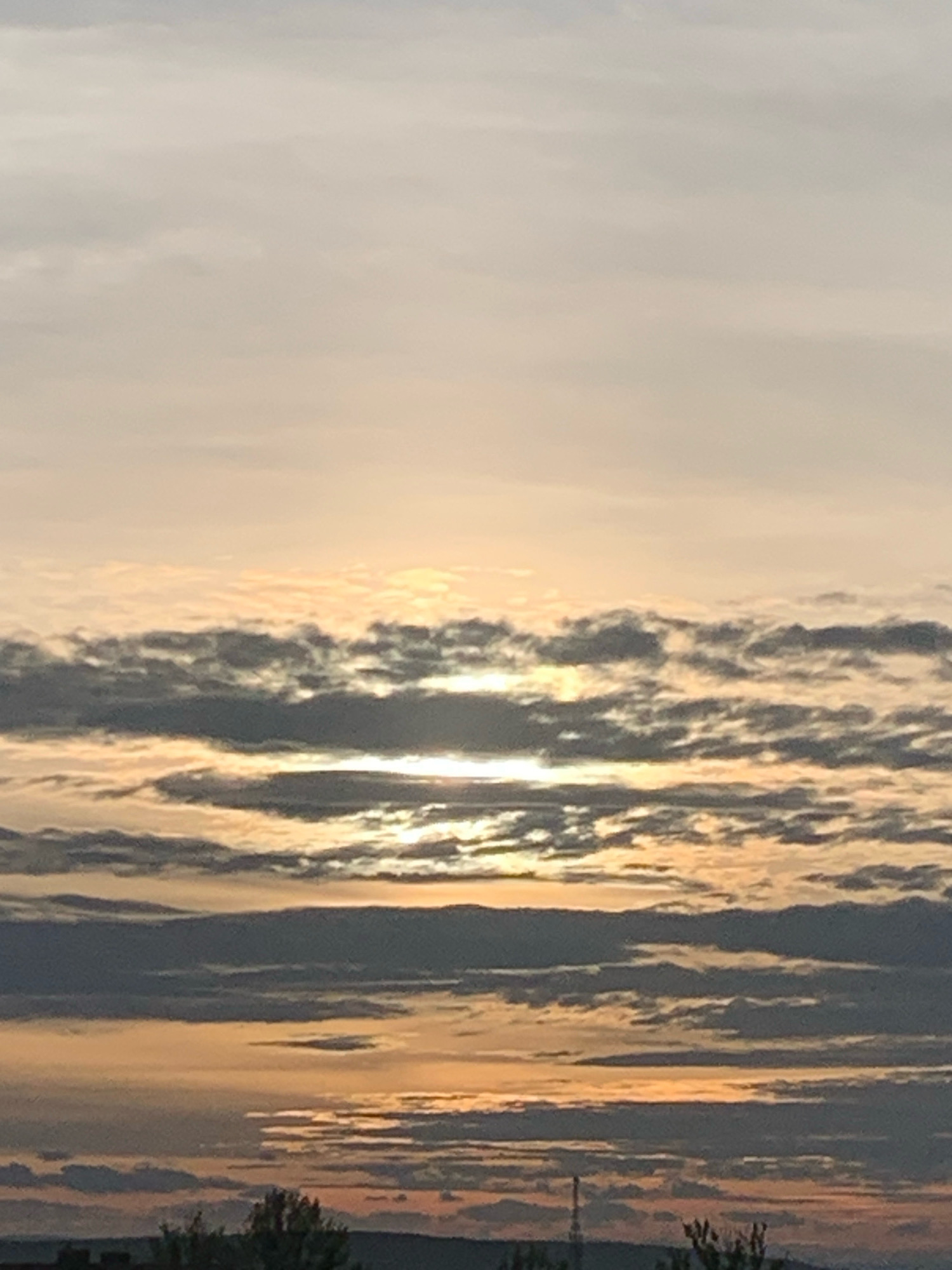 The image depicts a sky filled with layered clouds at sunset, casting a warm, orange glow. The sun is partially obscured by the clouds, creating bright horizontal streaks of light. Below the clouds, the silhouette of a city skyline is visible, with trees and buildings forming a dark, indistinct band against the horizon. The overall scene is one of a tranquil evening sky over an urban landscape. The location in the city of Kolomyia, Ukraine is not discernible from the image.