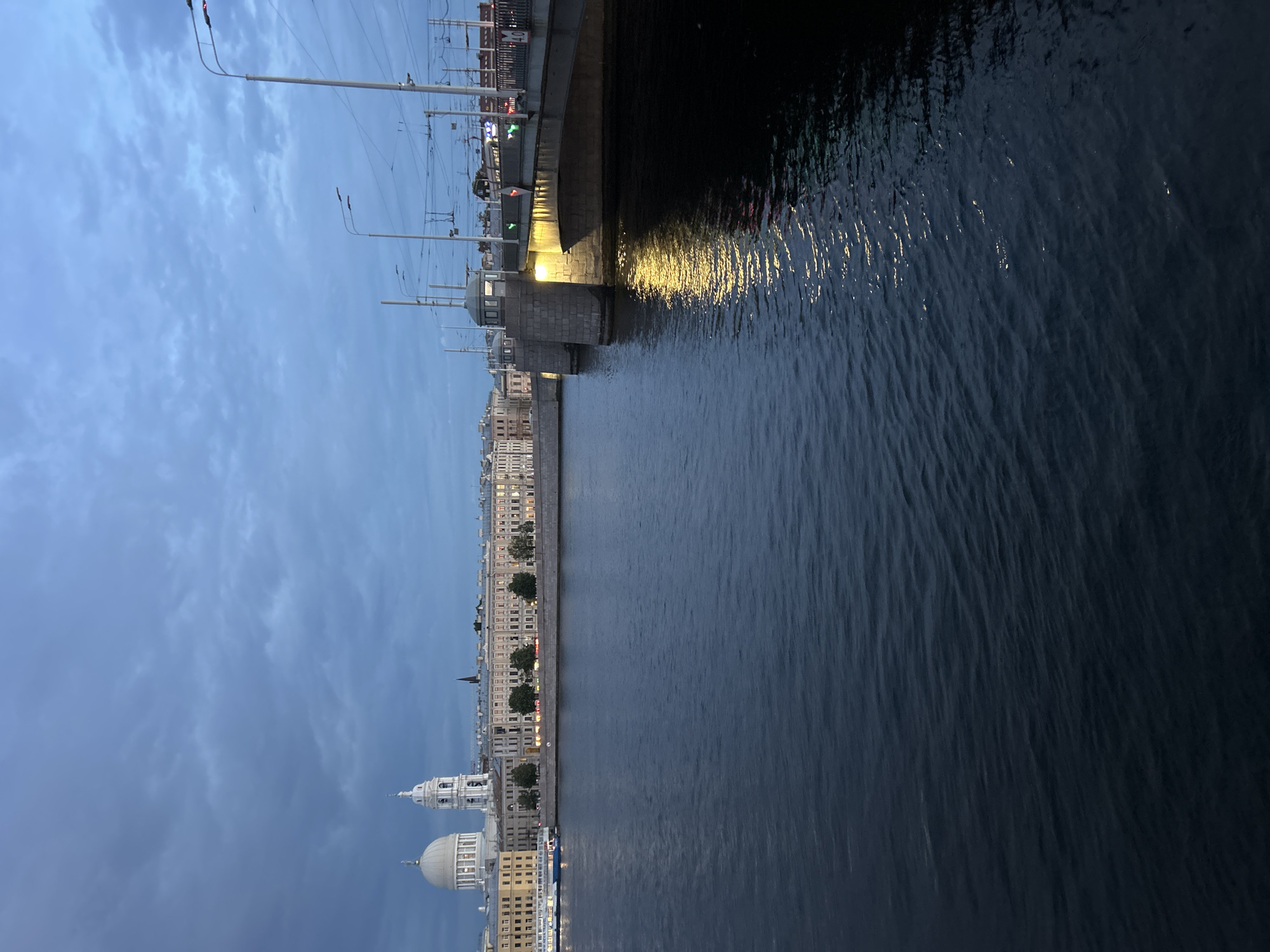 An urban scene shows a body of water in the foreground, with a bridge and buildings on the far bank. The sky is overcast and a deep blue. The water is dark and choppy, with reflections of lights from the bridge and buildings. The bridge is made of stone and concrete, with streetlights and tram wires overhead. The buildings are a mix of architectural styles, with some featuring domes and classical detailing. A boat is visible on the water near the far bank. The overall mood is quiet and somewhat somber, suggesting dusk or early evening.