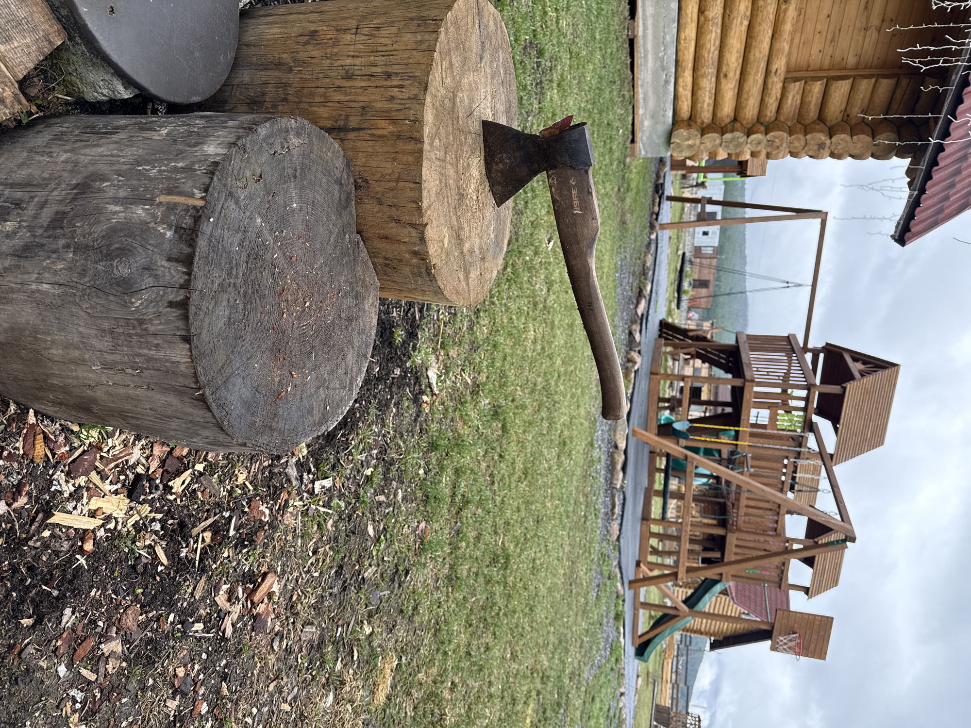 This image depicts a rustic outdoor scene, likely a yard or recreational area in Tyulyuk, Russia. In the immediate foreground, two large, horizontally placed wooden logs rest on the ground. The log closest to the viewer is dark grey with prominent growth rings, while the one behind it is a lighter, warmer brown. A dark, circular metal object is partially visible behind the darker log.
An axe is conspicuously stuck into the top surface of the lighter brown log. It features a dark, worn metal head and a wooden handle, which bears the faint inscription "396E". The ground surrounding the logs is a mixture of dark soil, wood chips, and patches of green grass, suggesting recent wood-chopping activity.
Further back, a substantial multi-level wooden playground structure occupies the right side of the frame. This playset includes various platforms, railings, at least two slides (one green, one reddish-brown), and swings suspended by yellow chains. A small basketball hoop with a red net is attached to one part of the structure. To the upper right, a section of a traditional log cabin is visible, constructed with round, interlocking logs and featuring a red corrugated roof decorated with white string lights.
The sky is uniformly overcast and grey, indicating a cloudy daytime. In the distant background, beyond the playground, some faint structures and misty hills are visible under the dull light. No people are present, lending a quiet atmosphere to the scene.