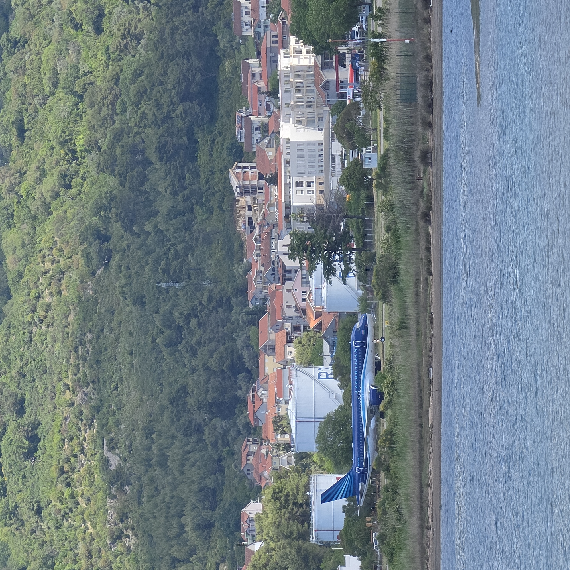 The image shows the coastal village of Đuraševići, Montenegro, viewed from an elevated position. A dense forest covers the hills to the left. A row of buildings, primarily residential with red-tiled roofs, lines the coastline. Modern, taller buildings are interspersed among the traditional structures. To the right, a calm body of water stretches towards the horizon. An aircraft, painted in blue and white with "Azerbaijan" written on it, is parked on the shoreline, near what appear to be large storage tanks. The vegetation between the buildings and the water's edge consists of low-lying grasses and shrubs. The lighting suggests it is daytime.