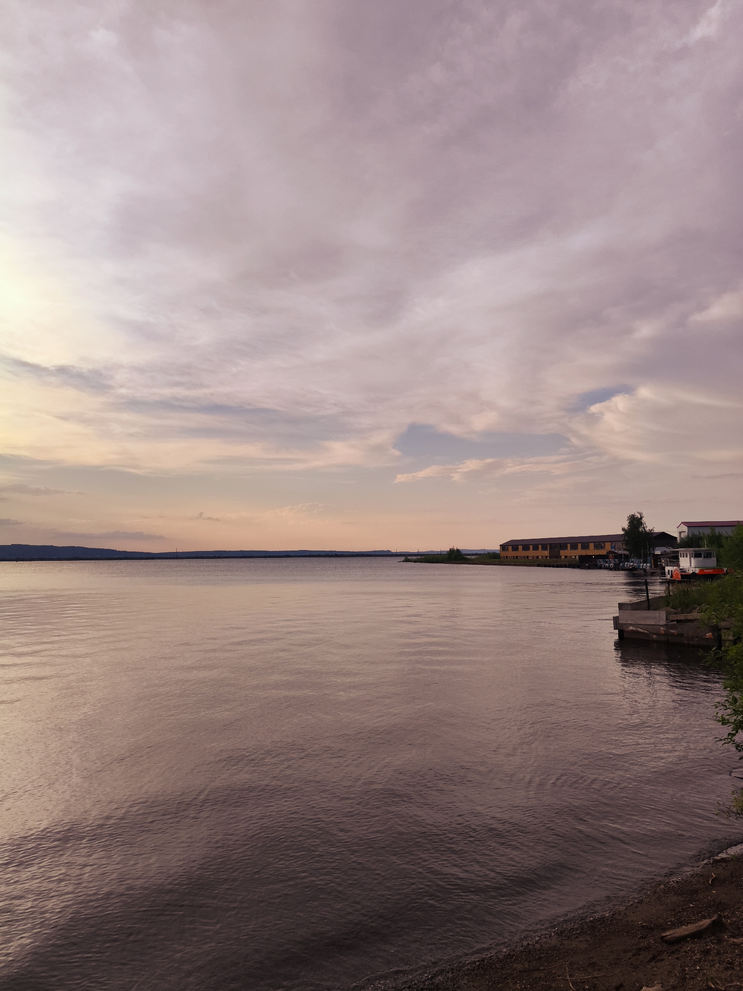 This is a vertical, eye-level, full shot of a wide, calm body of water, likely a river, with a distant shoreline and a cloudy sky at sunset or sunrise. The water reflects the soft, muted colors of the sky, creating a tranquil atmosphere.
On the right side of the frame, a small wooden dock juts into the water, with a white boat tied to it. Beyond the dock, a line of buildings, including a long, yellow, industrial-looking structure and a smaller red-roofed building, are visible along the shoreline. Green trees and foliage are also present. The shoreline on the left is distant and appears hilly or mountainous, creating a dark silhouette against the sky.
The sky is filled with wispy, scattered clouds, tinged with shades of pink, purple, and pale yellow, indicative of either sunrise or sunset. The overall mood is peaceful and serene. There is no visible text in the image. The location context is Chaikovsky, Russia.