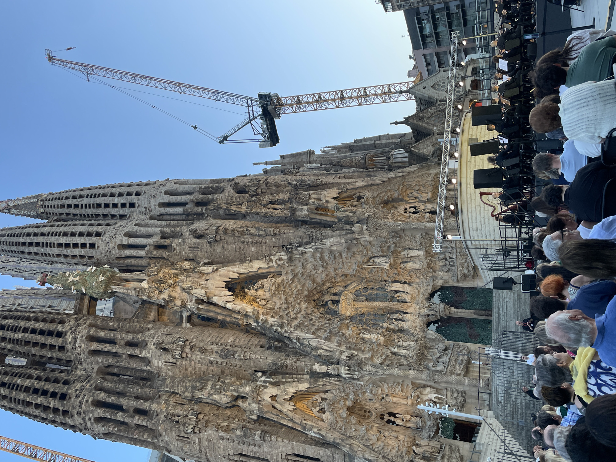 The image depicts an outdoor concert taking place in front of the Sagrada Familia basilica in Barcelona, Spain. An orchestra is performing on a stage set up on a plaza, with a large audience seated in front of them. The Sagrada Familia, an iconic unfinished Roman Catholic minor basilica, dominates the background with its intricate facade and towering spires, one of which is under construction with a crane visible. The clear blue sky suggests it is daytime. The audience members are diverse in age and are seated on chairs, facing the stage. The overall scene captures a cultural event set against a world-renowned architectural landmark.