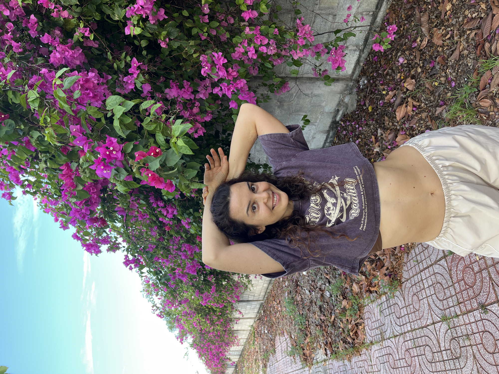 A woman with curly brown hair stands outdoors in front of a wall covered in vibrant pink bougainvillea flowers. She is wearing a dark purple cropped t-shirt and light-colored loose-fitting pants. Her arms are raised with her hands behind her head, and she is smiling at the camera. The ground in front of the wall is paved with patterned tiles and covered in fallen leaves and dry grass. The background shows a blue sky with scattered clouds. This scene is located on a walkway in Phu Thuy Ward, Vietnam.