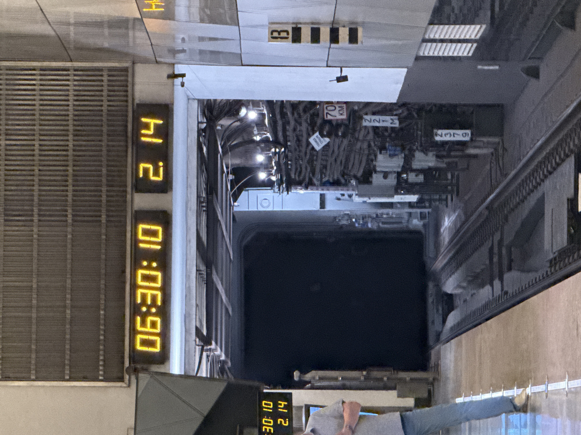 The image depicts a metro station in Moscow, Russia. A digital clock displays 06:30:10 and 2.14. A person in a light-colored shirt and blue jeans stands in the foreground. A train tunnel stretches into the distance. Overhead signs indicate location and distance, including "70 km" and other alphanumeric codes like "2376" and "2-14." The station has metal grills and a platform edge with tactile warning strips. Platform number '13' is visible. Overhead electrical cables are visible.