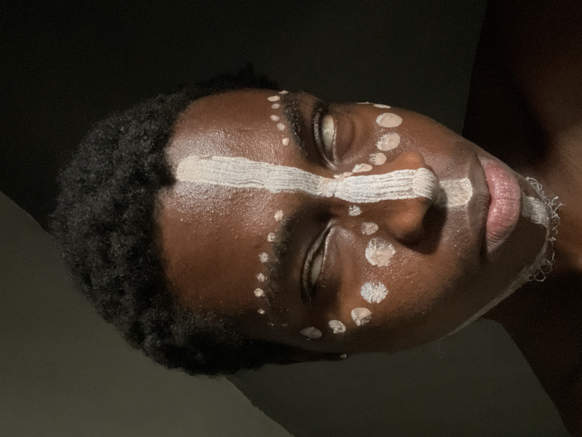 A close-up, medium-shot photograph depicts a person with dark skin, likely of African descent, lying on their side. Their eyes are closed. White paint adorns their face in a stripe across the forehead and a series of dots along the face. The lighting is dramatic, with a dark background and shadows highlighting the facial features. The photograph was not taken in Onitsha, Nigeria.