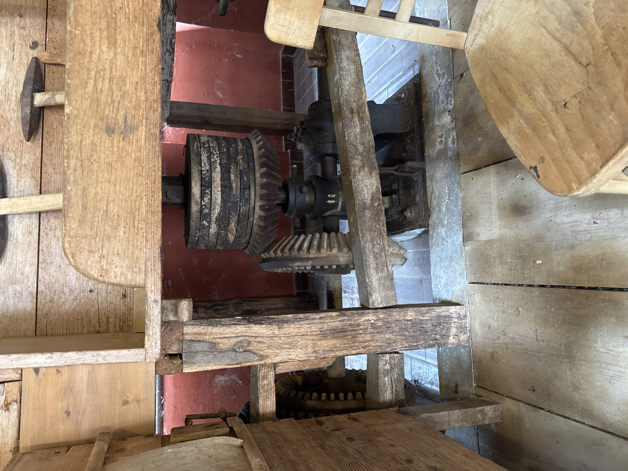 The image shows a close-up, eye-level view of a vintage, large-scale, wooden and metal grinding mechanism, likely part of a historic mill or workshop, located inside a building with red walls and a tiled floor. Dominating the center is a large, stacked set of heavy, dark-colored discs forming a weight or grindstone, with a prominent toothed gear wheel below it. This gear meshes with another large wooden gear to its left and a smaller metal gear to its right. The mechanism is supported by robust wooden beams and posts, some appearing worn and aged. To the right foreground, a light-colored wooden chair is partially visible, its seat facing away from the camera. The overall scene suggests an industrial or agricultural heritage setting, potentially within the Postojna Cave complex in Postojna, Slovenia, which historically features various machinery and displays related to local industries and traditions. The lighting is somewhat dim, with natural light possibly entering from the left, casting subtle shadows. The condition of the machinery suggests it is either a preserved exhibit or a functional piece of antique equipment.