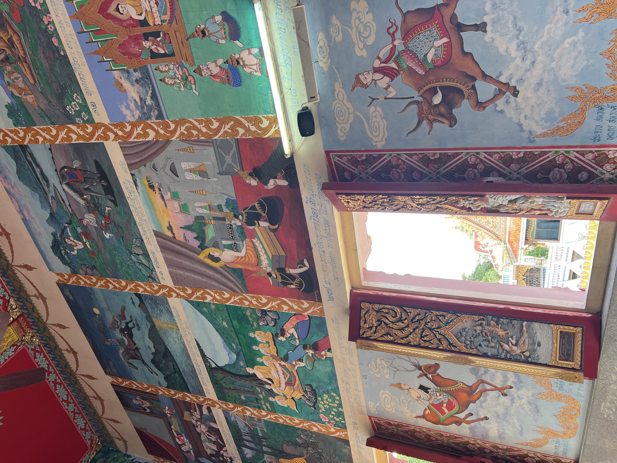 The image depicts a mural on the wall of the Baan Plai Laem Temple in Koh Samui, Thailand. The mural is divided into several panels, each featuring a scene with religious or mythological figures. The style is traditional Thai art, characterized by vibrant colors and detailed illustrations.
In the upper sections, the panels show figures on horseback, a woman in labor, and a procession of people. One panel depicts a seated Buddha figure surrounded by monks and devotees. Another panel illustrates a man riding a bull through the clouds.
The lower part of the mural, visible through an open doorway, shows a figure riding a deer. The doorway itself is elaborately decorated with floral patterns and gold leaf. In the background, through the doorway, parts of the temple architecture and its surroundings are visible. The overall impression is one of a richly decorated religious site, likely a temple or sanctuary.