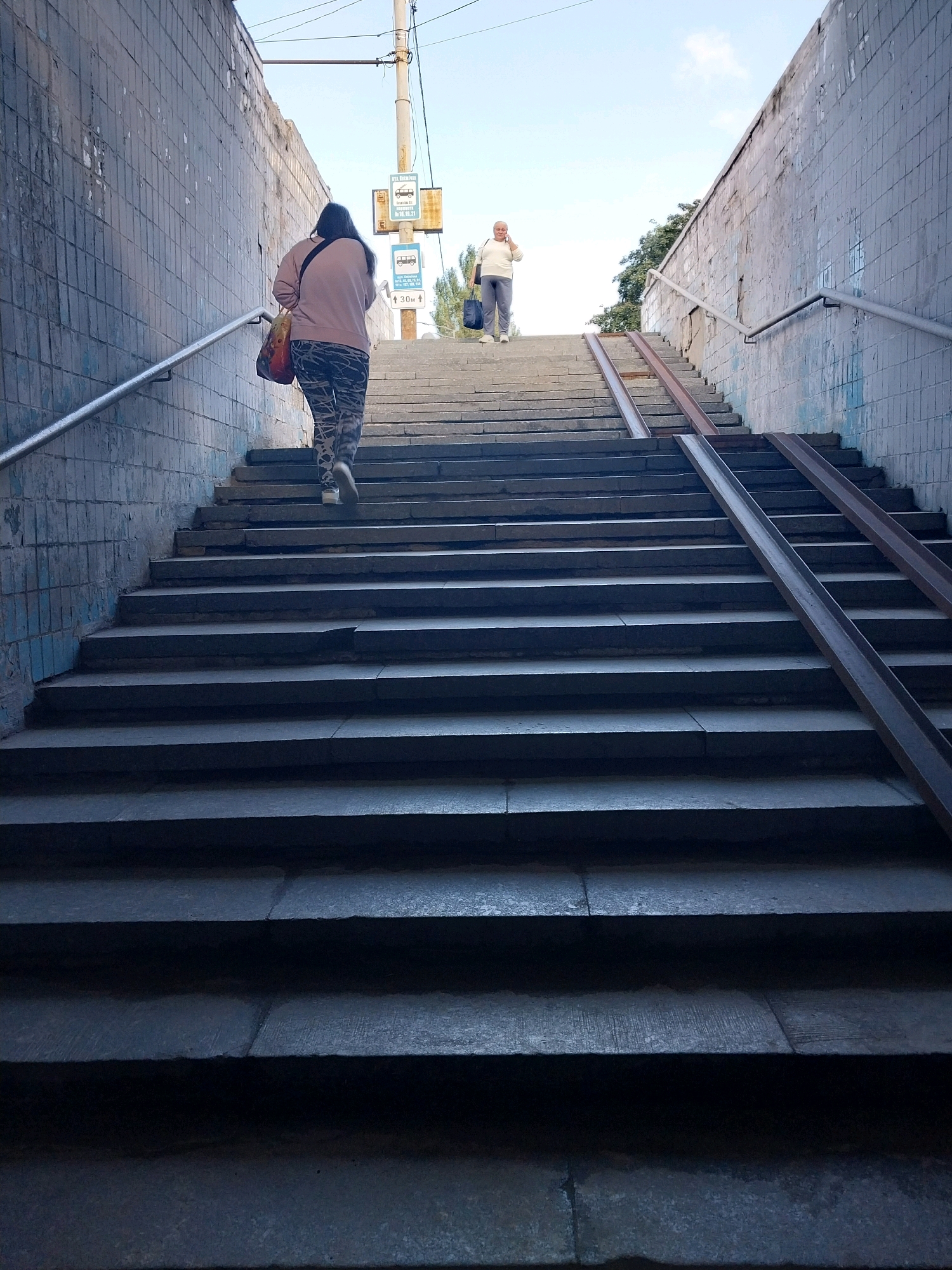 The image depicts an outdoor staircase in an urban environment, likely Dnipro, Ukraine, given the signage. Two individuals are ascending the steps. In the foreground, a person wearing patterned leggings and a light pink sweatshirt walks up the stairs, carrying a multi-colored bag over their shoulder. Further up the staircase, another person, dressed in white and grey, is walking and holding a dark blue bag. The staircase is made of concrete, with metal railings on either side. A metal ramp runs alongside a section of the stairs. The walls flanking the staircase are tiled and show signs of age and weathering. Overhead, a utility pole with street signs is visible against a blue sky with scattered clouds. The overall impression is of a public access point, possibly an underpass or an elevated walkway, in a moderately maintained urban setting.