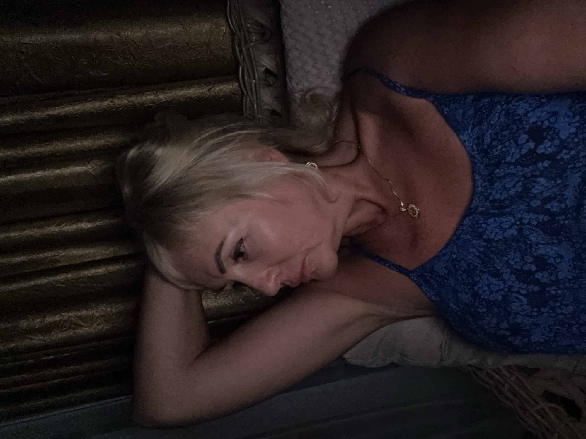 A medium shot captures a blonde woman from the chest up, reclining on her left side with her left arm behind her head. She is wearing a blue floral-patterned dress and a delicate gold necklace with a pendant. The woman has fair skin and is looking off to her left with a contemplative expression. Her hair is styled in a loose, tousled manner. Behind her, a textured, dark brown curtain provides a backdrop, with a hint of a woven headboard or furniture visible to the right. The lighting is dim, casting shadows that emphasize the contours of her face and body. The image appears to be a self-portrait, taken indoors in what could be a bedroom or living area in Alanya, Turkey, possibly during the evening or in low-light conditions.