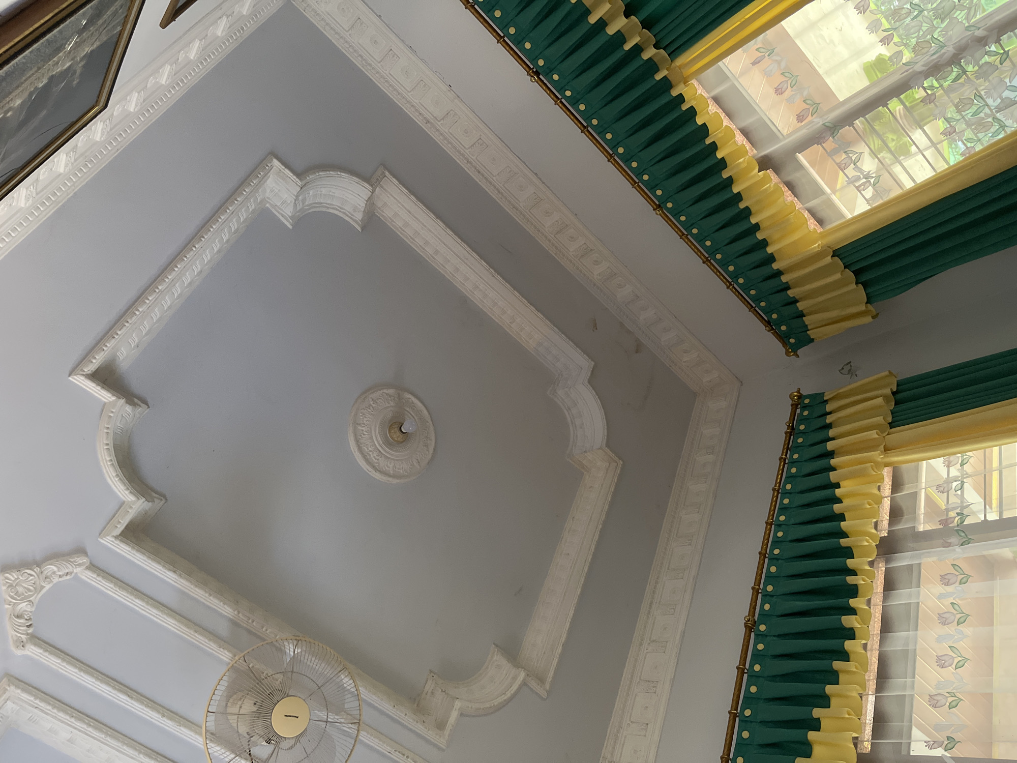 The image shows a view of an ornate white ceiling with intricate moldings and a central light fixture. A ceiling fan is visible in the upper left corner. The ceiling appears to be in a residential interior. The image is taken looking upwards from within a room. Decorative curtains in green and yellow are visible along the bottom of the frame, covering a window. The lighting suggests an indoor environment. The location of Glagah Kulon, Indonesia, is provided context but is not directly observable in the image itself.