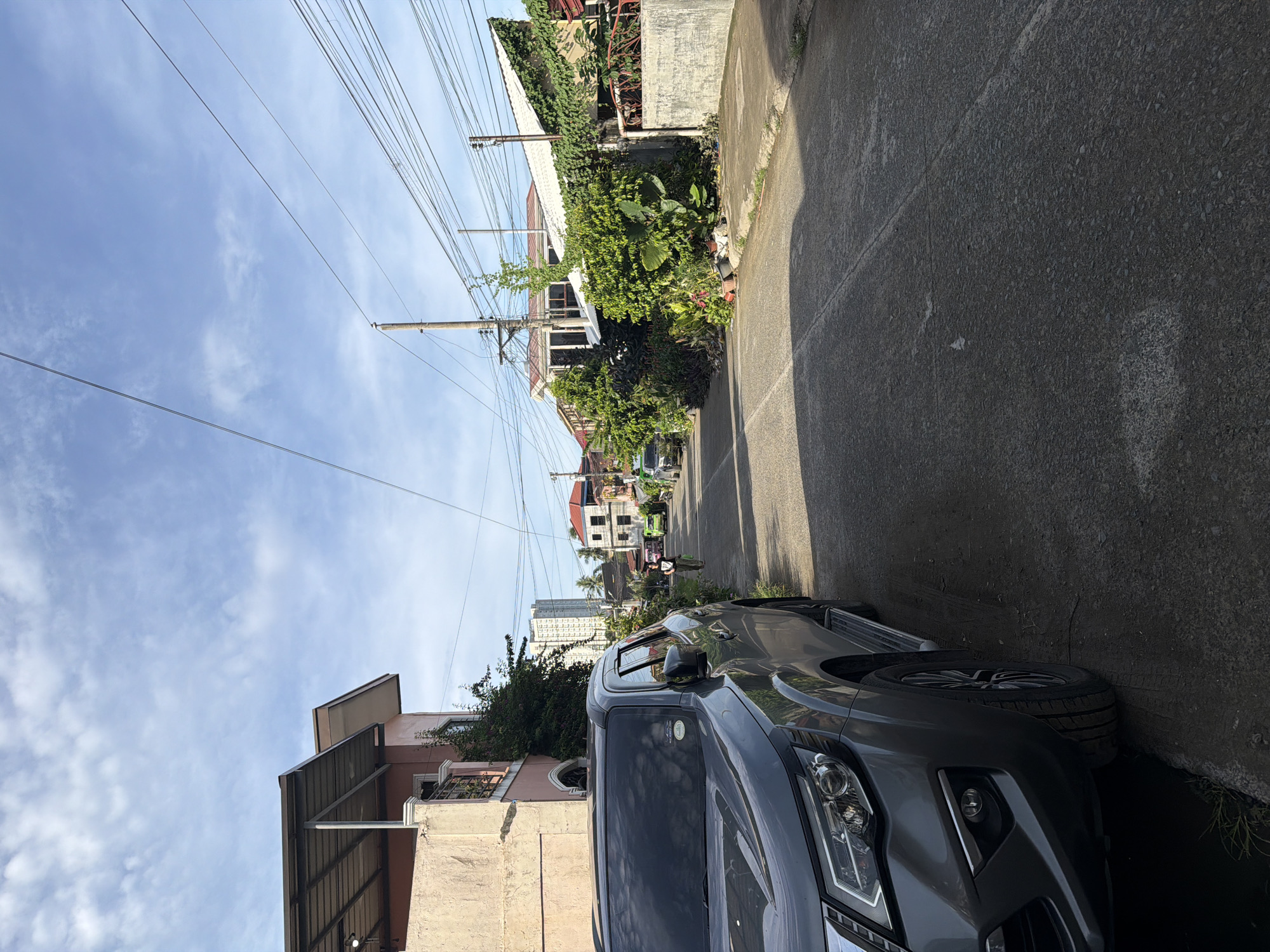 The image depicts a residential street in Davao City, Philippines. A gray SUV is parked on the right side of the road. Power lines stretch across the sky. The street is lined with houses and vegetation. A person on a motorcycle is visible further down the street, with other vehicles also present. In the distance, a tall building is visible. The weather appears sunny.