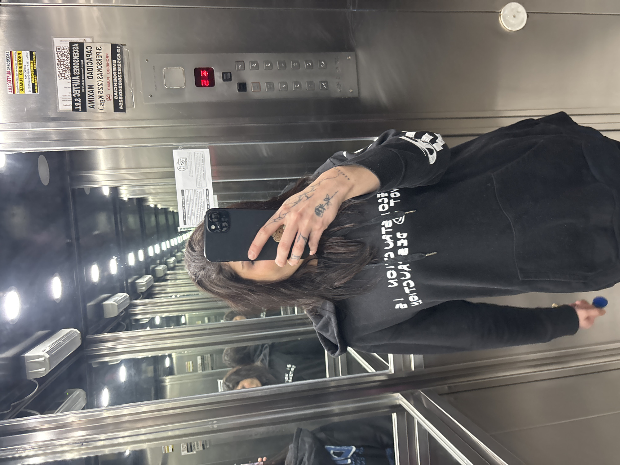 A person takes a selfie in an elevator with stainless steel walls, reflecting multiple images of the person in a repeating pattern. The person is wearing a black hooded sweatshirt with white lettering. A smartphone obscures part of the person's face. On the wall are elevator control panels and various signage, including emergency instructions in Spanish. The number "21" is illuminated on the elevator floor indicator. Due to the limited information and visual data, I cannot provide details about the city of Buenos Aires or specific actions, interactions, or more forensic details.