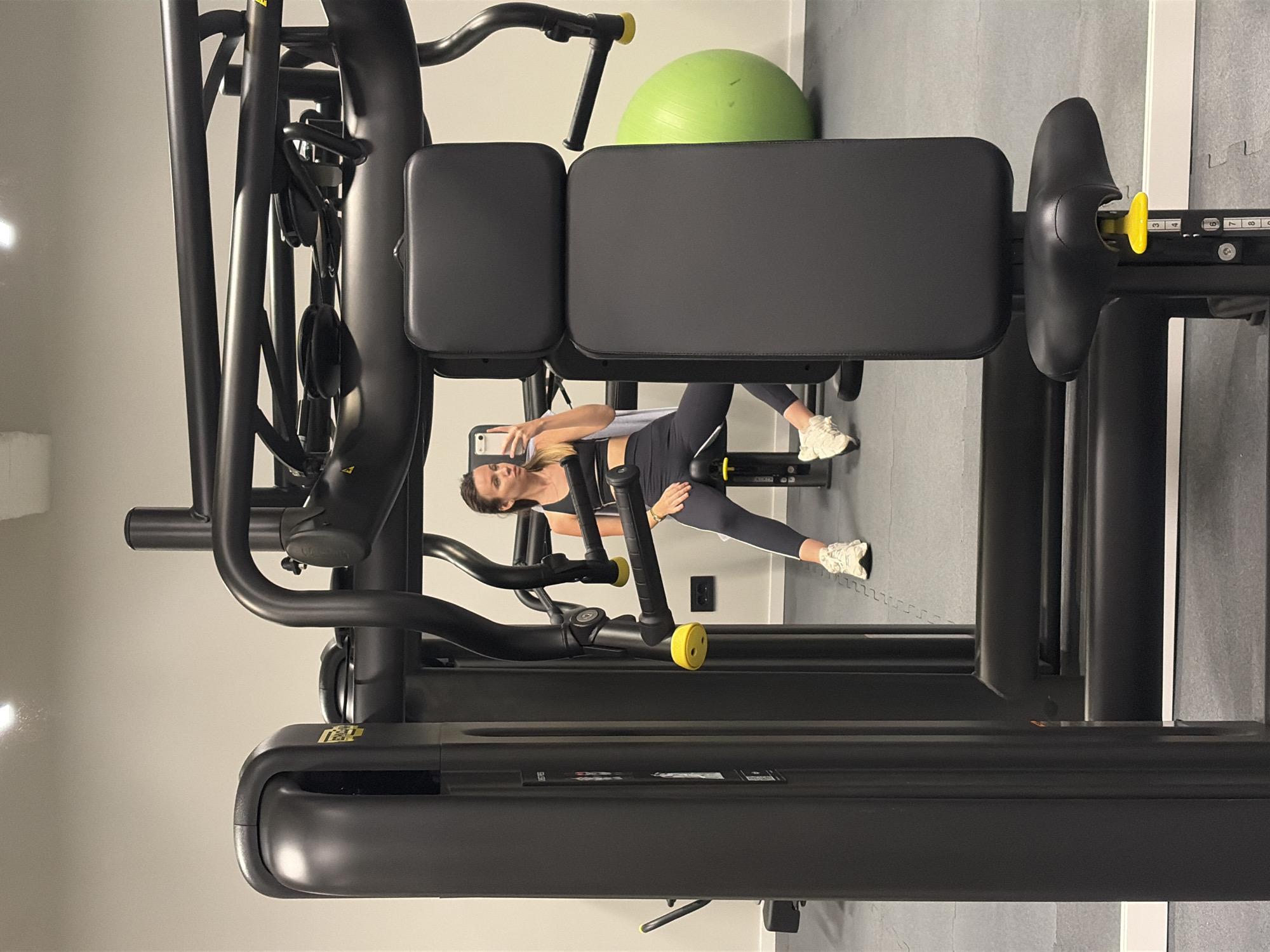 A woman in a black sports bra and black leggings is sitting on a black weight machine, holding a cell phone in her left hand, as if taking a selfie. The weight machine is in a gym with a green exercise ball in the background, near a gray wall and a gray floor.
