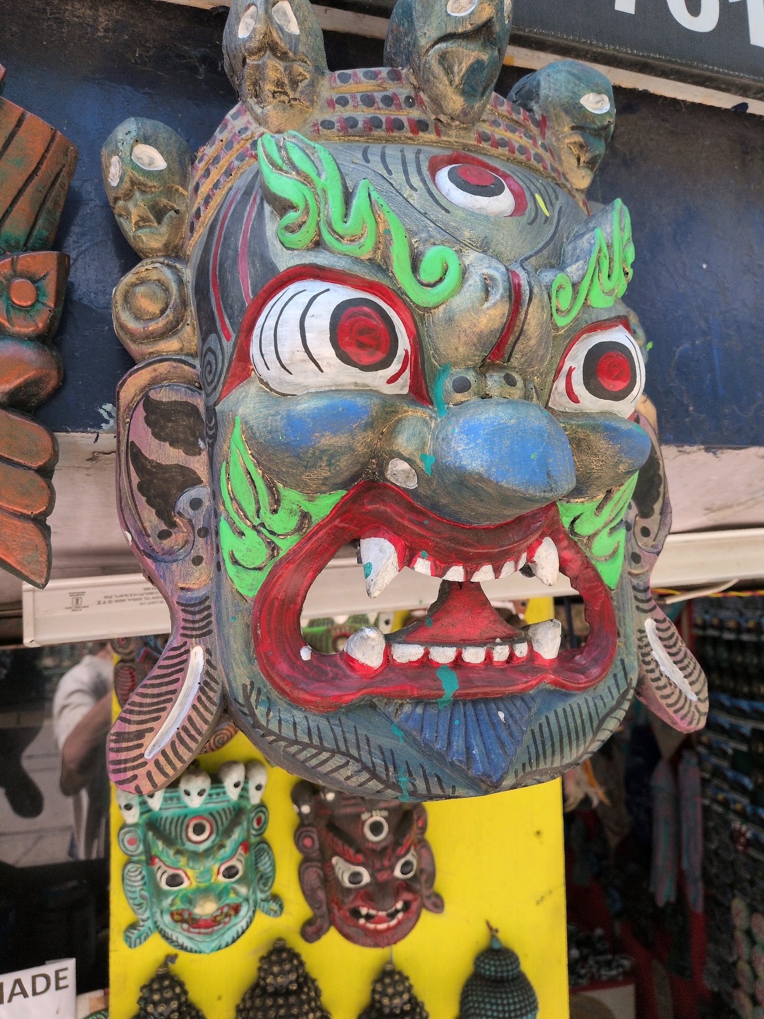 This is a close-up, eye-level, outdoor, full-frame photograph of a collection of colorful, carved wooden masks displayed for sale in Bhagsu, India. The central focus is a large, ornate mask with a fearsome expression, painted in shades of gray, red, green, blue, and white. It features wide, staring eyes with red pupils, a gaping mouth with sharp white teeth and a red tongue, and green decorative elements around the forehead and cheeks. Flanking this central mask are smaller, less detailed masks. To the left, a partially visible mask is carved from wood with brown and orange tones. Below the central mask, on a bright yellow background, are two smaller, more traditional-looking masks: one predominantly green with three eyes, and another dark red with two eyes. Scattered at the bottom are dark, textured decorative elements that appear to be part of the display. In the background, out of focus, are hints of a street market with hanging fabrics and other goods. The lighting suggests it is daytime, and the overall impression is of a vibrant, cultural display of local crafts.