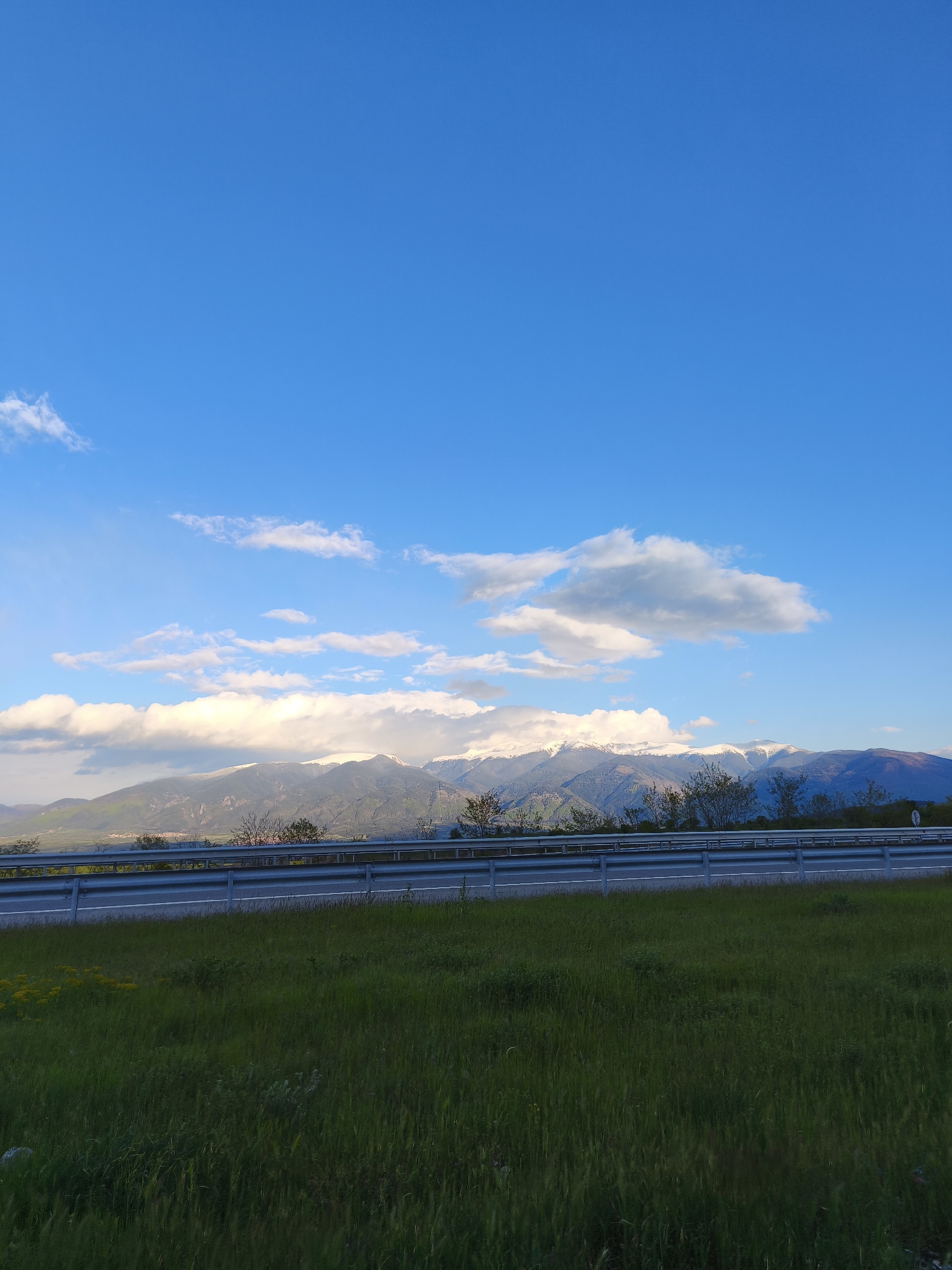The image depicts a landscape with a vibrant blue sky occupying the upper half of the frame, dotted with fluffy white clouds. A mountain range stretches across the midground, their peaks partially covered with snow. In the foreground, a grassy field extends towards the viewer, with a metal guardrail separating it from what appears to be a road. Based on the location provided, this scene could be near Blatino, Bulgaria. There are no people visible in the image.